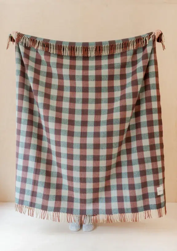 A person stands behind a large, fringed checkered blanket in muted blue and brown tones, with only their feet visible below the blanket's edge.