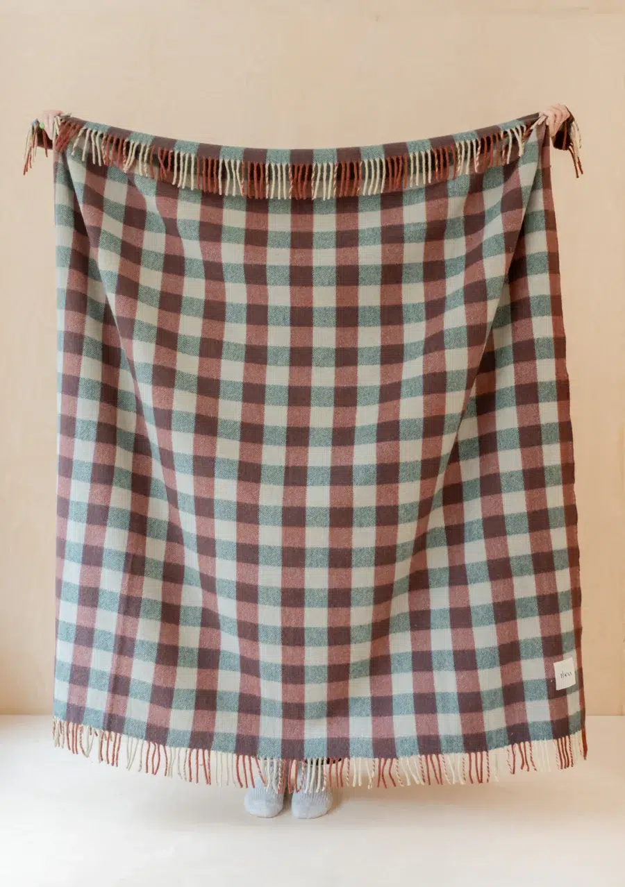 A person stands behind a large, fringed checkered blanket in muted blue and brown tones, with only their feet visible below the blanket's edge.