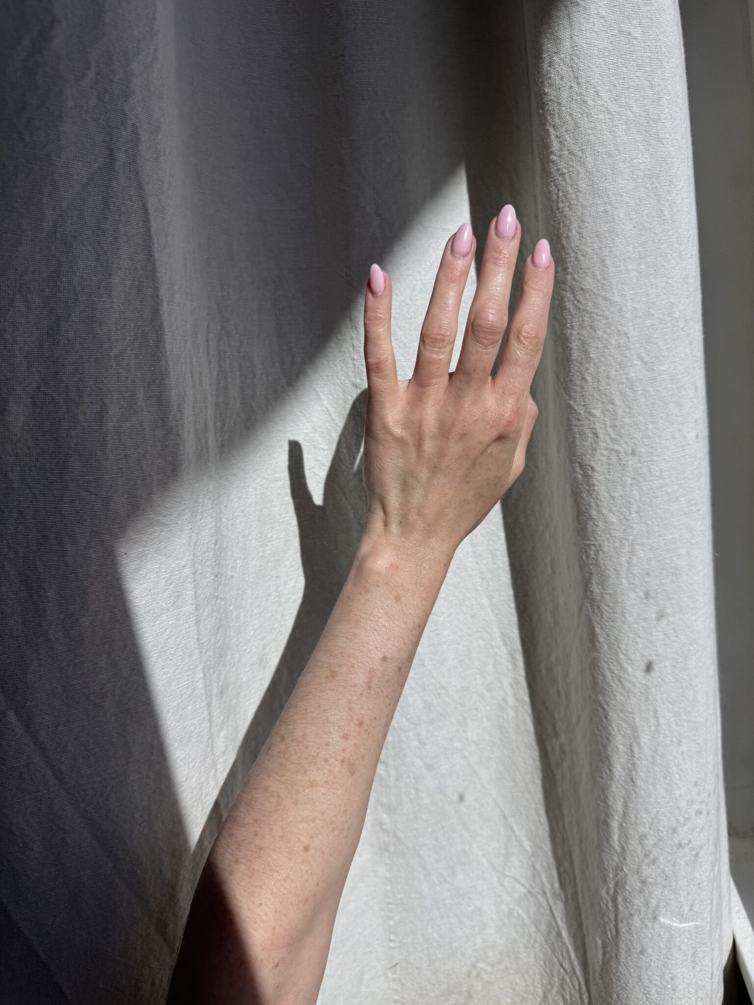 A hand with light pink nails is raised toward a white curtain, casting a shadow on the fabric in bright sunlight.