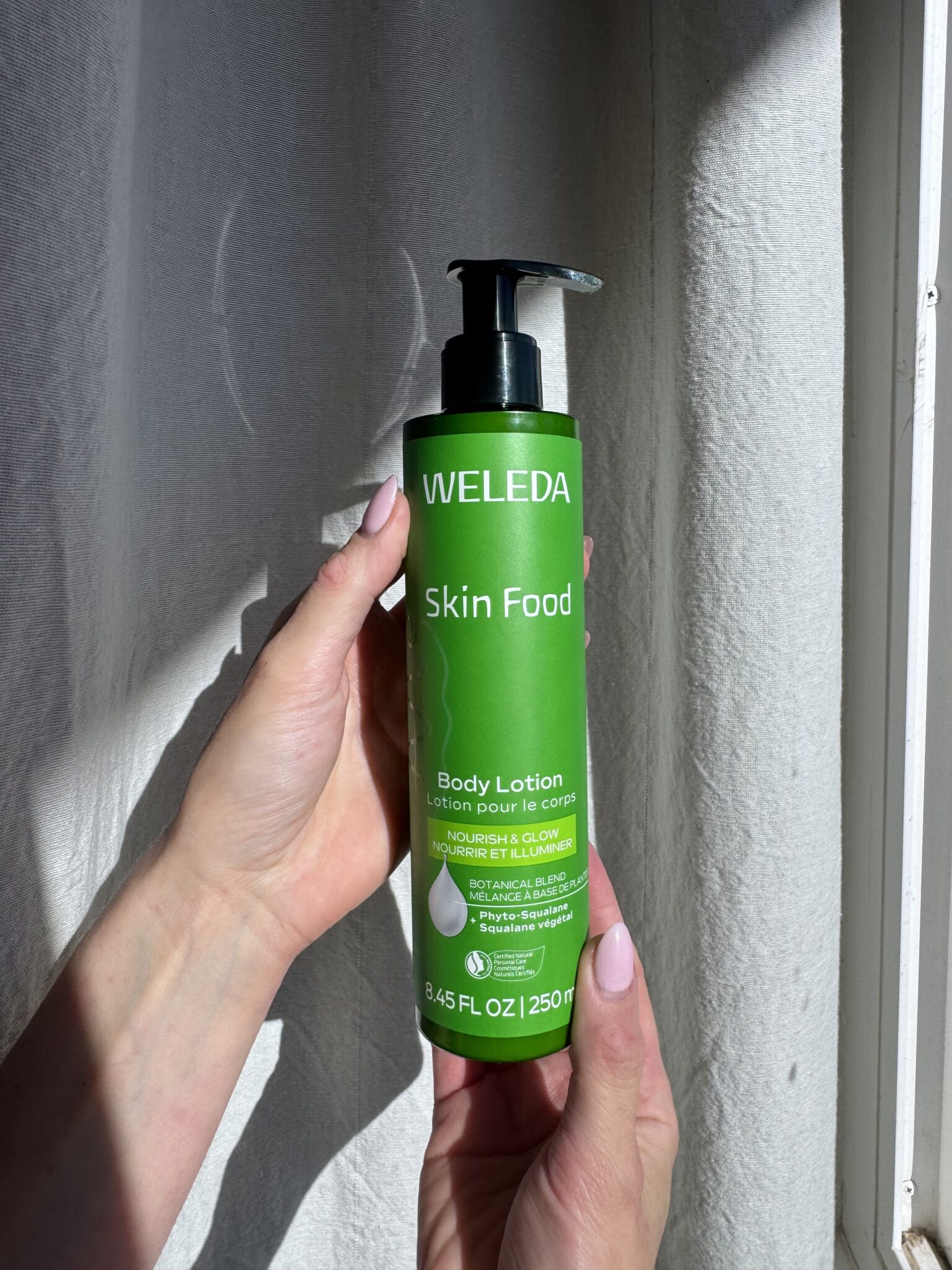 A person holds a green bottle of Weleda Skin Food Body Lotion, 8.45 fl oz, with a pump dispenser, in natural light by a white curtain.