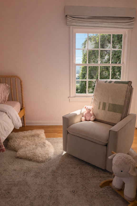 A beige armchair with a pink teddy bear sits near a sunlit window in a child's bedroom. A small bed, plush rug, and stuffed animal are also visible.