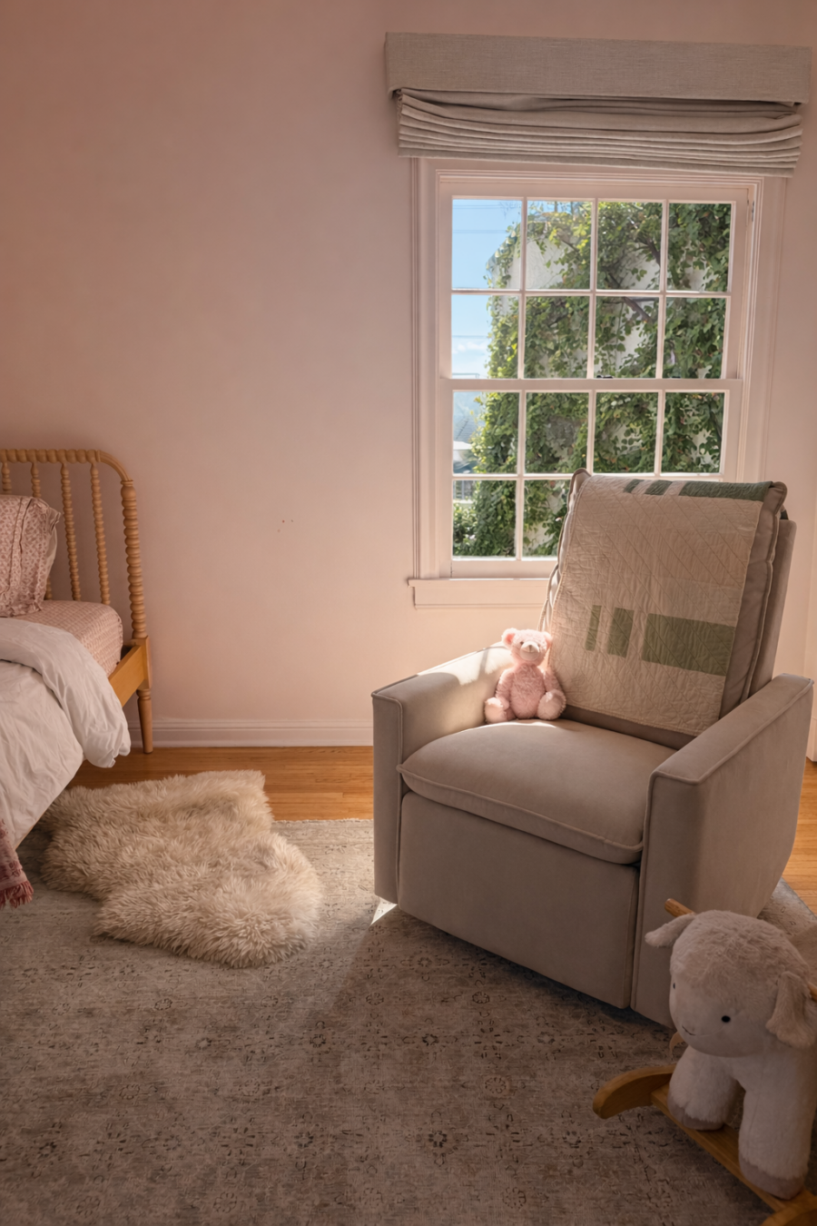 A beige armchair with a pink teddy bear sits near a sunlit window in a child's bedroom. A small bed, plush rug, and stuffed animal are also visible.