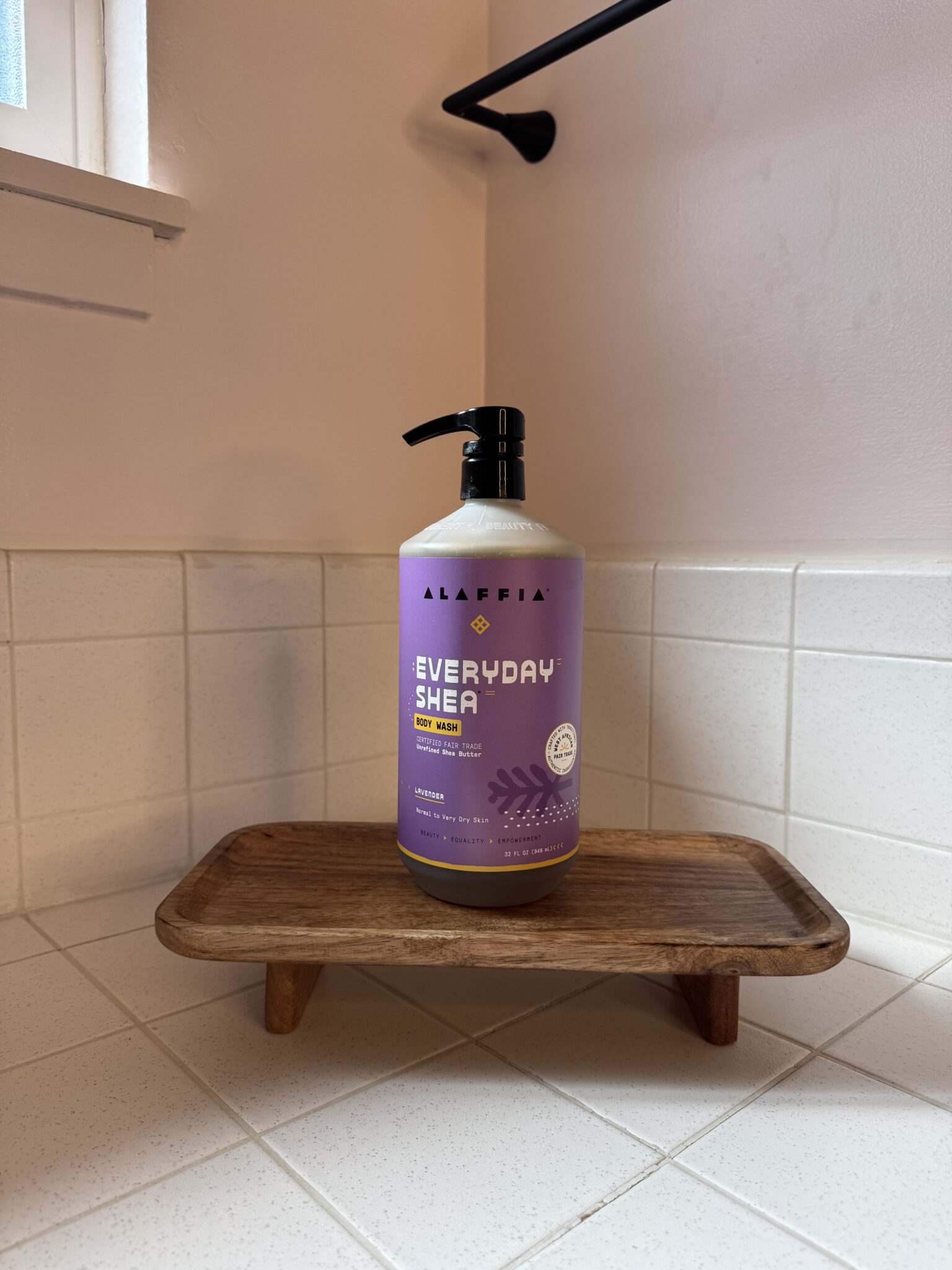 A bottle of Alaffia Everyday Shea body wash with a pump stands on a small wooden tray in a tiled bathroom corner.