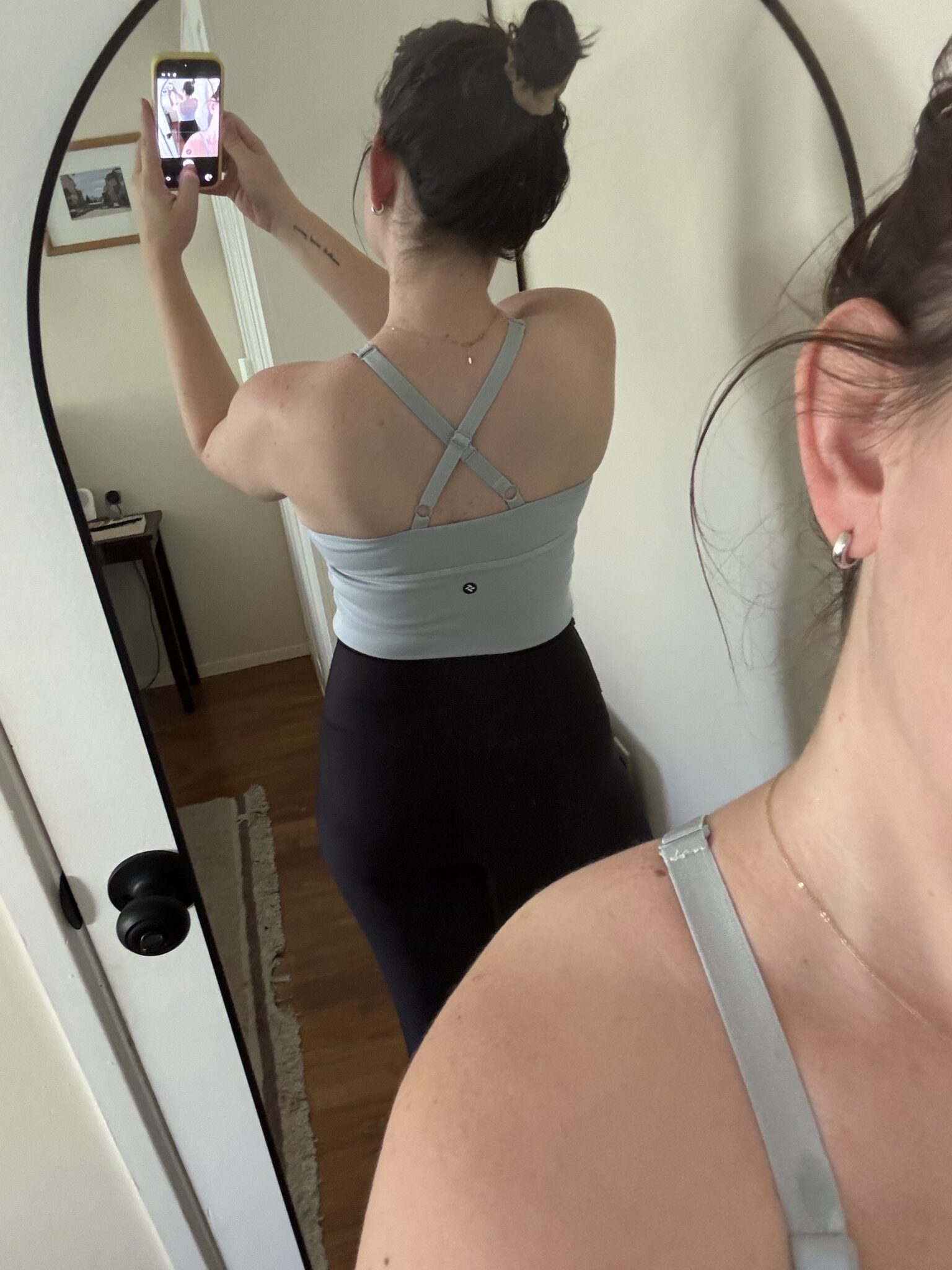 A woman stands in front of a mirror, taking a photo of her back and shoulders in a light blue strappy top and black pants; a partial view of her face is in the foreground.