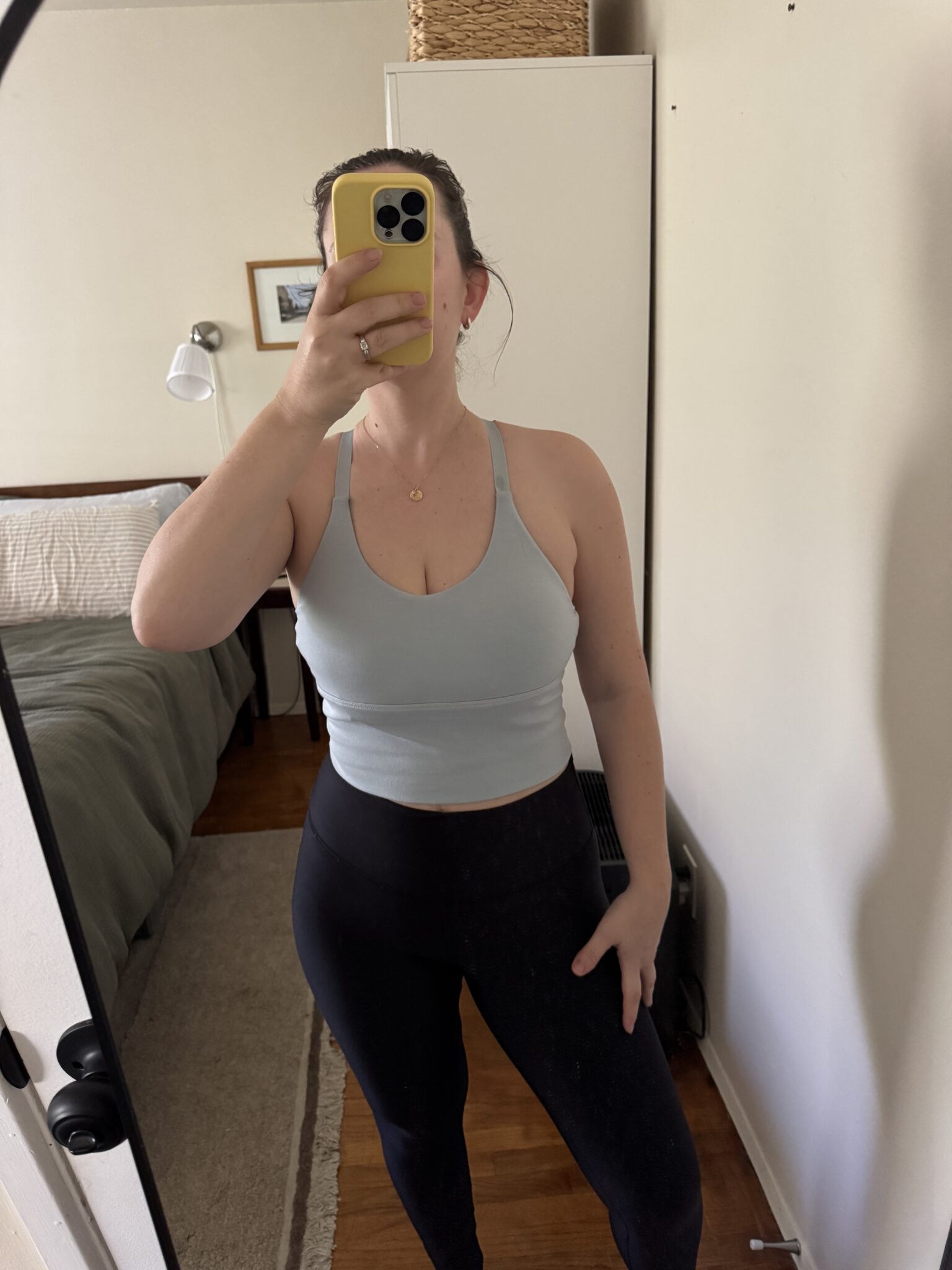 A person takes a mirror selfie in a bedroom, wearing a light blue sports bra and black leggings, with their face partially covered by a yellow phone.
