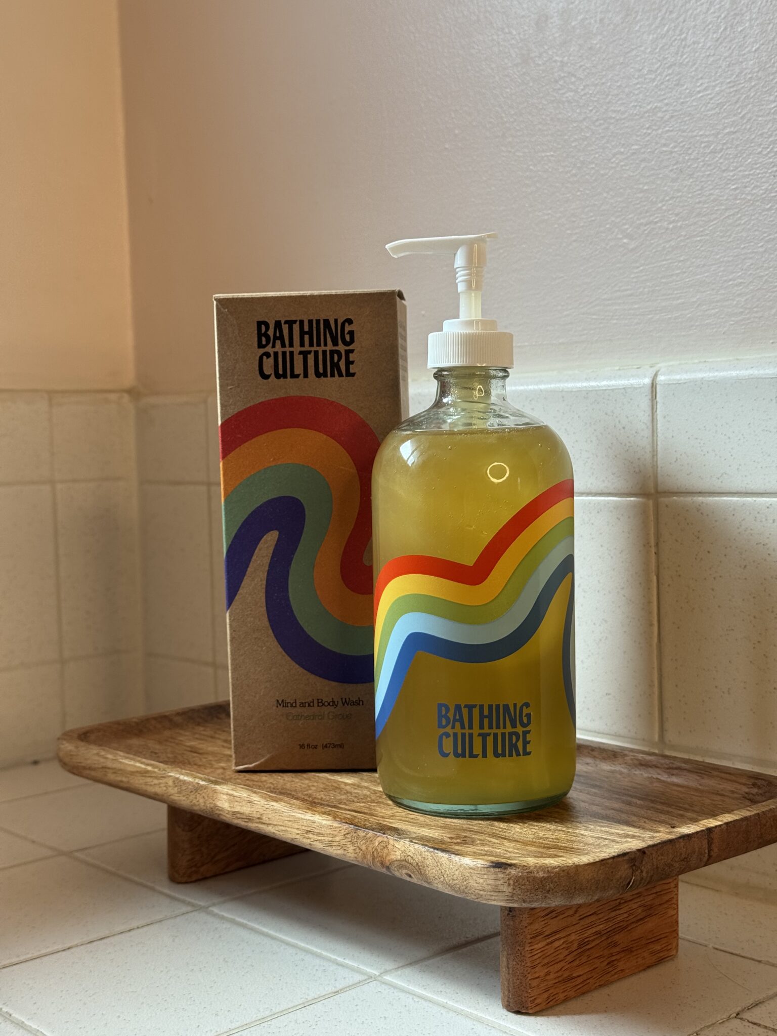 A glass pump bottle and box of Bathing Culture body wash with colorful wavy stripes, placed on a wooden tray on a tiled bathroom counter.