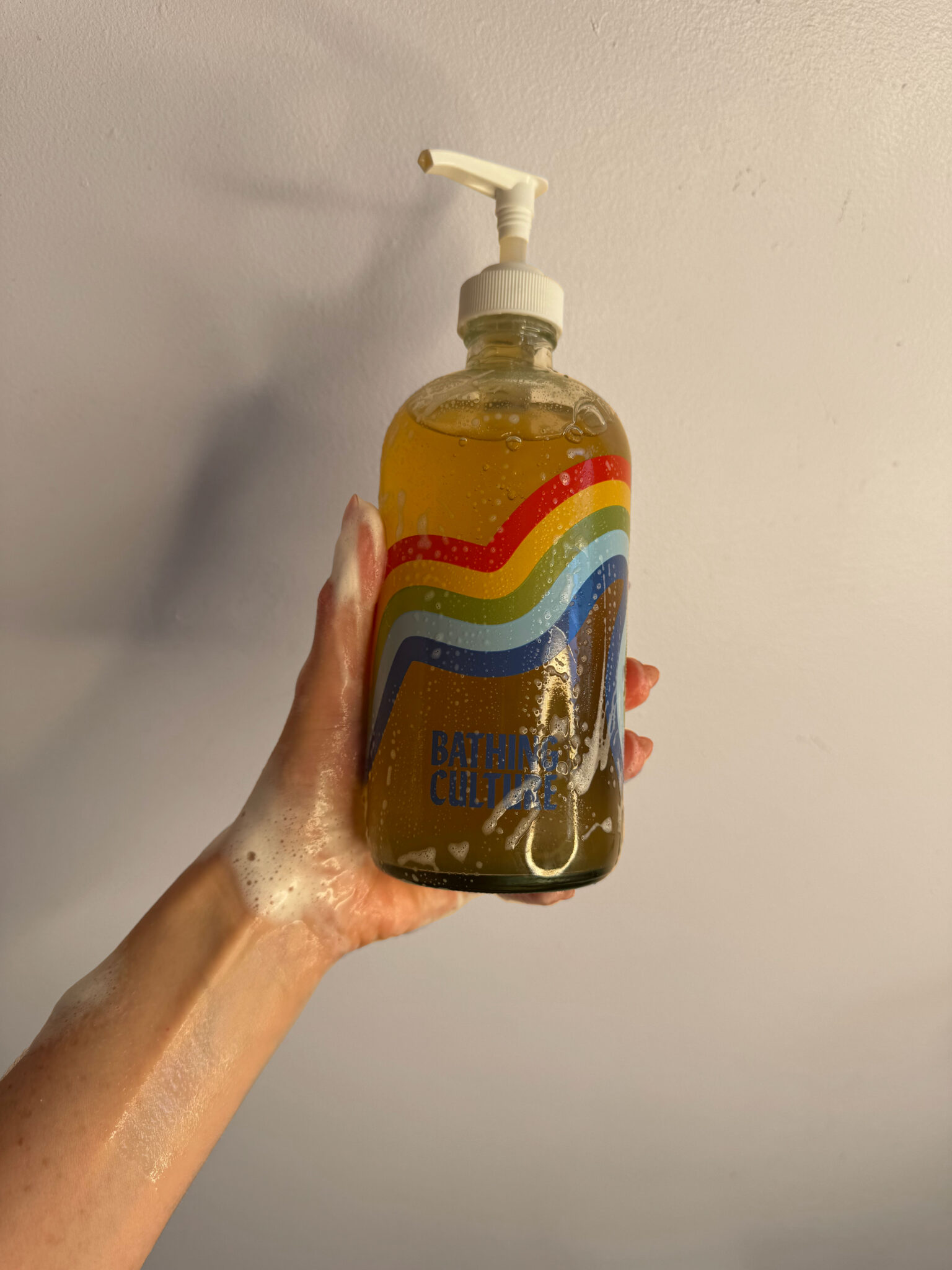 A hand covered in soap holds up a clear pump bottle of Bathing Culture body wash with a rainbow design on the label against a plain light background.