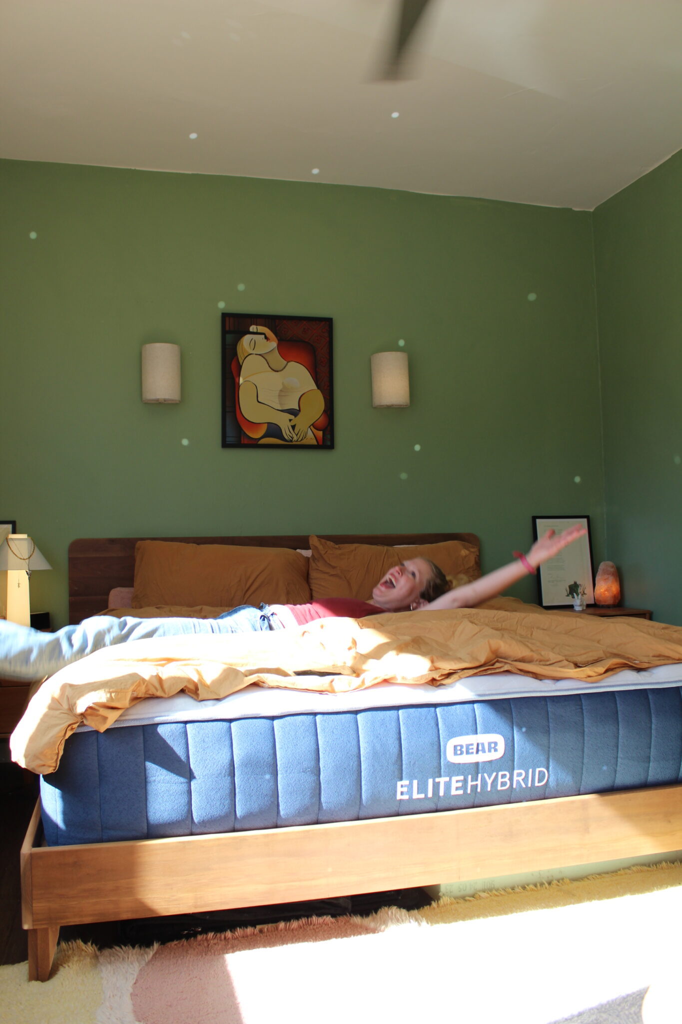 A person lies on a bed with arms outstretched in a sunlit bedroom. The bed features an organic Bear Elite Hybrid mattress, and the room showcases green walls and modern decor.