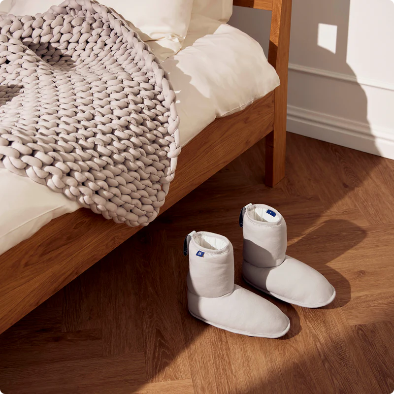 A pair of cozy slippers rest on a wooden floor beside a bed adorned with a chunky knit blanket and crisp white bedding.