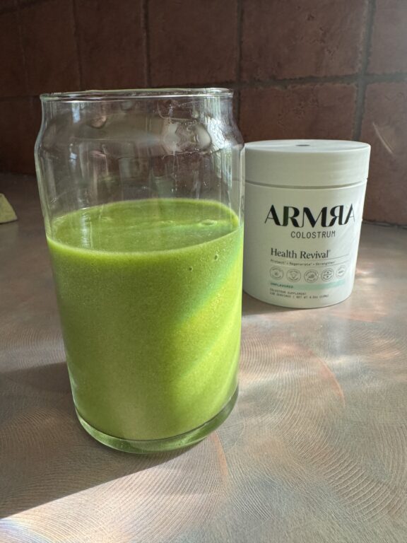 A glass filled with green liquid sits on a countertop beside a container labeled “ARMRA Colostrum Health Revival.”.