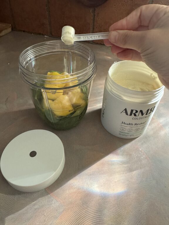 A hand adds a scoop of ARMRA Colostrum powder to a blender cup filled with leafy greens and pineapple chunks. The supplement container and lid are nearby.