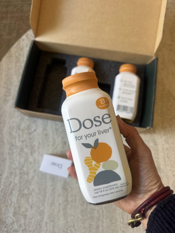 A hand holds a Dose for your liver supplement bottle with two more bottles and a small card in an open box on a table.