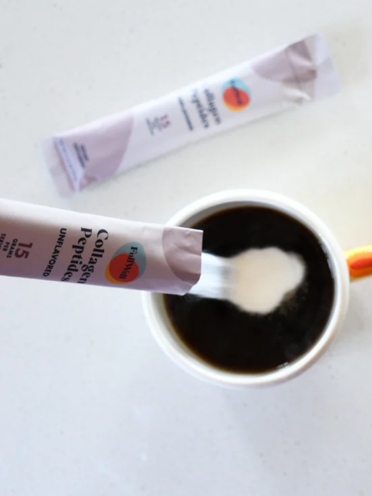 A packet of collagen peptides powder is being poured into a cup of black coffee on a white surface.