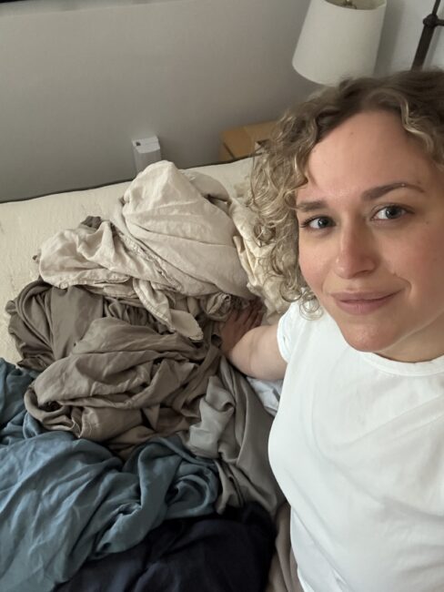 Person with curly hair wearing a white shirt sits on a bed next to a pile of unfolded laundry in neutral and blue tones.