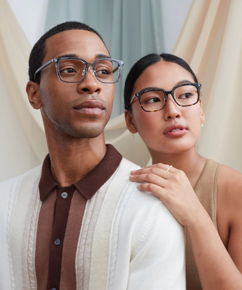 A man and a woman, both wearing glasses, stand close together indoors. The man looks forward while the woman looks slightly to the side, with her hand resting on his shoulder.