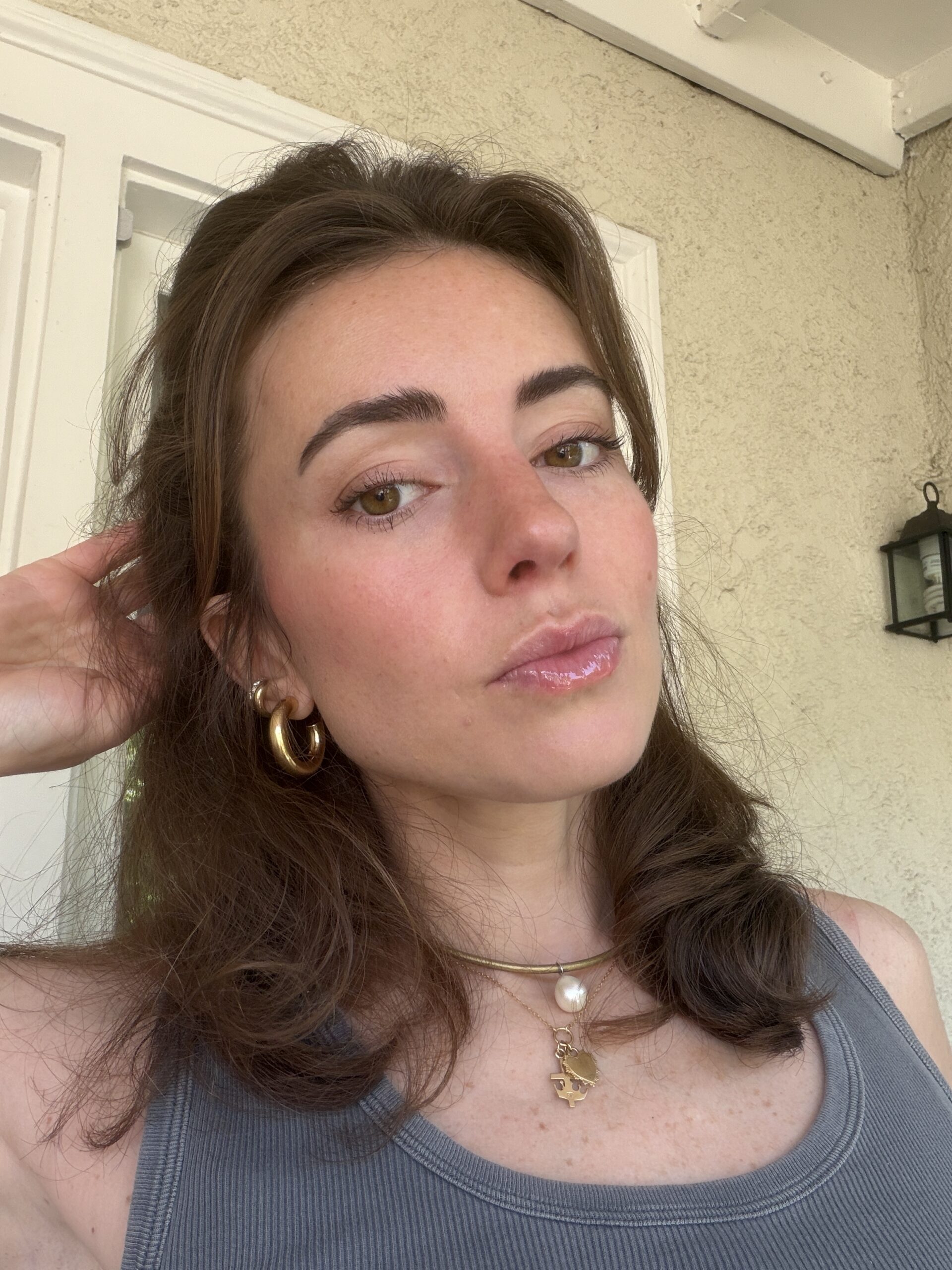 A woman with brown hair wearing a gray tank top, gold hoop earrings, and layered necklaces poses for a selfie in natural light. She stands in front of a light-colored wall and white door.