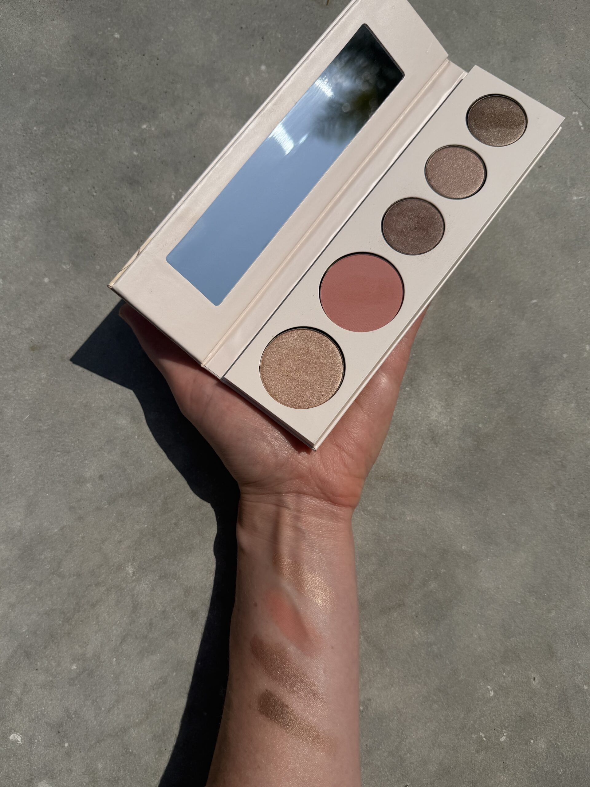 A hand holds an open makeup palette with a mirror, four eyeshadows, and one blush; swatches of the products are visible on the forearm.