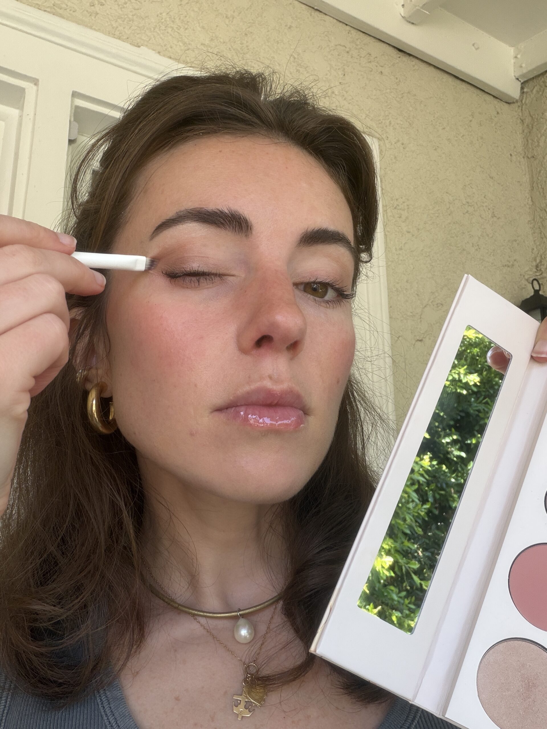 A woman applies eyeshadow with a brush to her eyelid while holding an open makeup palette with a mirror.