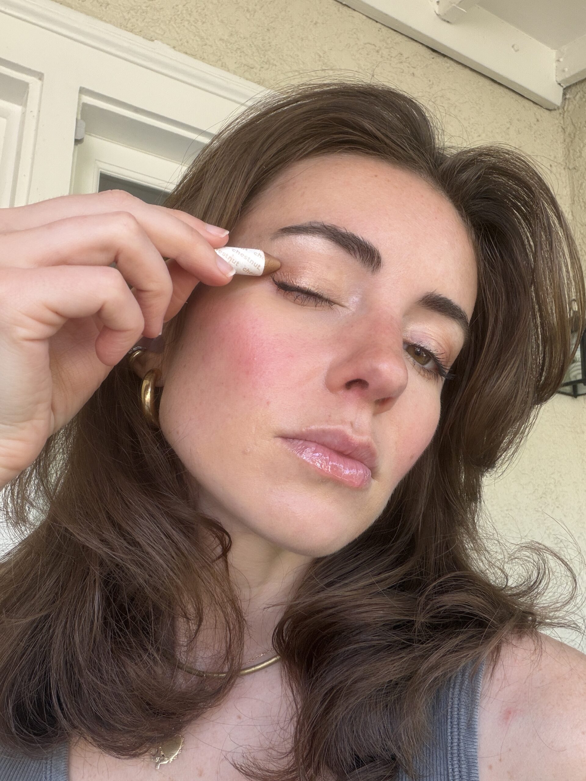 A woman applies shimmery eyeshadow to her eyelid using a makeup applicator, standing outdoors near a beige wall.