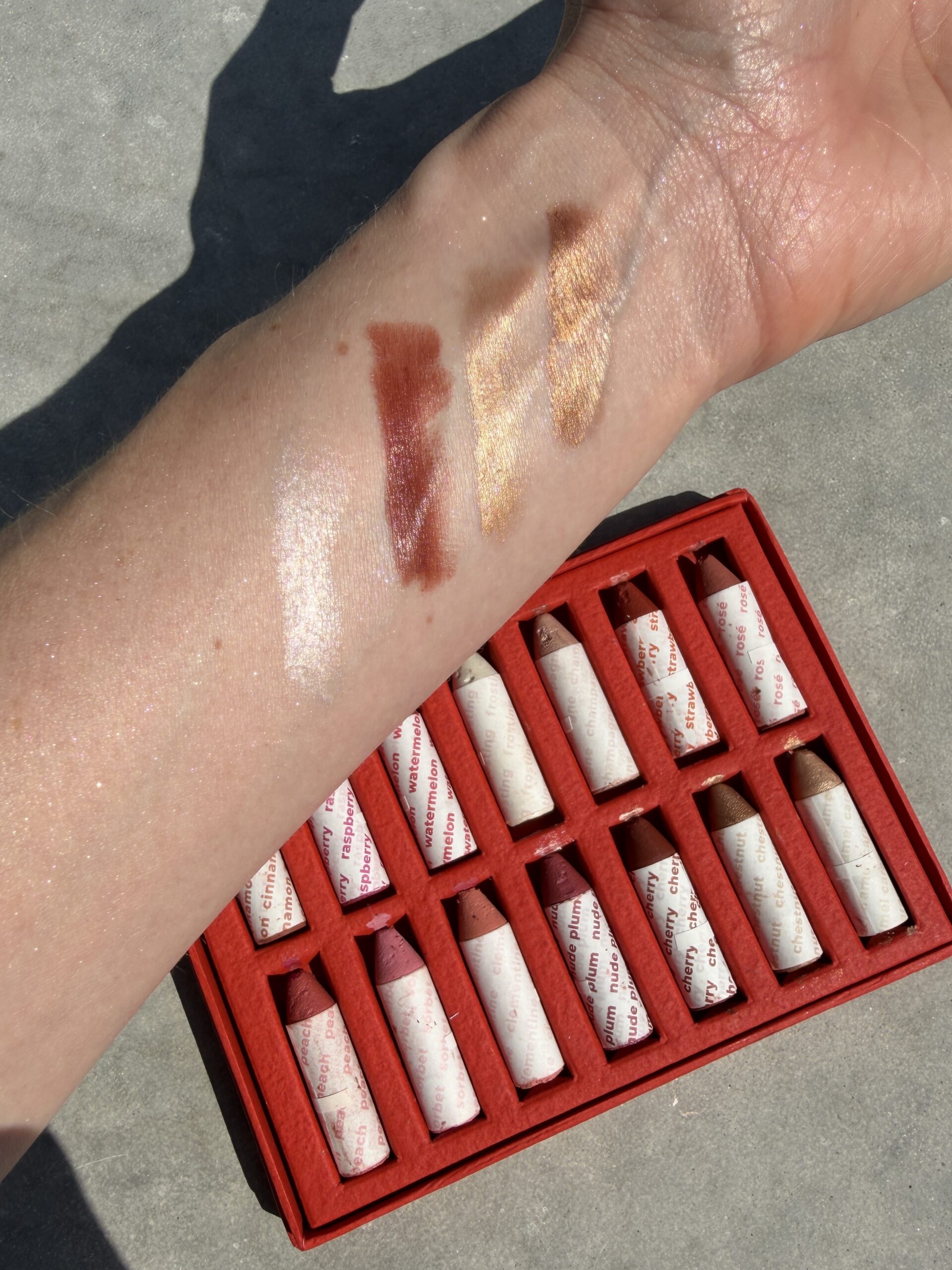 An arm displays three shimmery makeup swatches beside a red box containing 12 lipstick tubes arranged in two rows.
