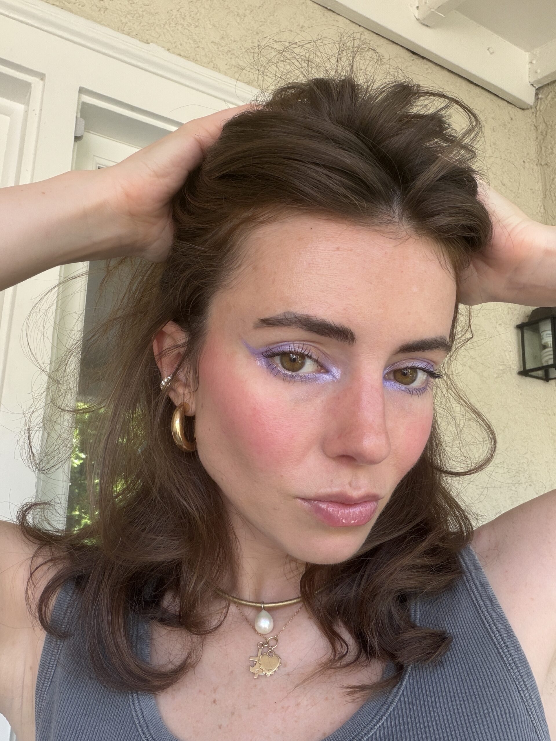 Woman with brown hair poses with hands on head, wearing light purple eyeshadow, blush, a grey tank top, gold hoop earrings, and layered necklaces, standing in front of a light-colored wall.