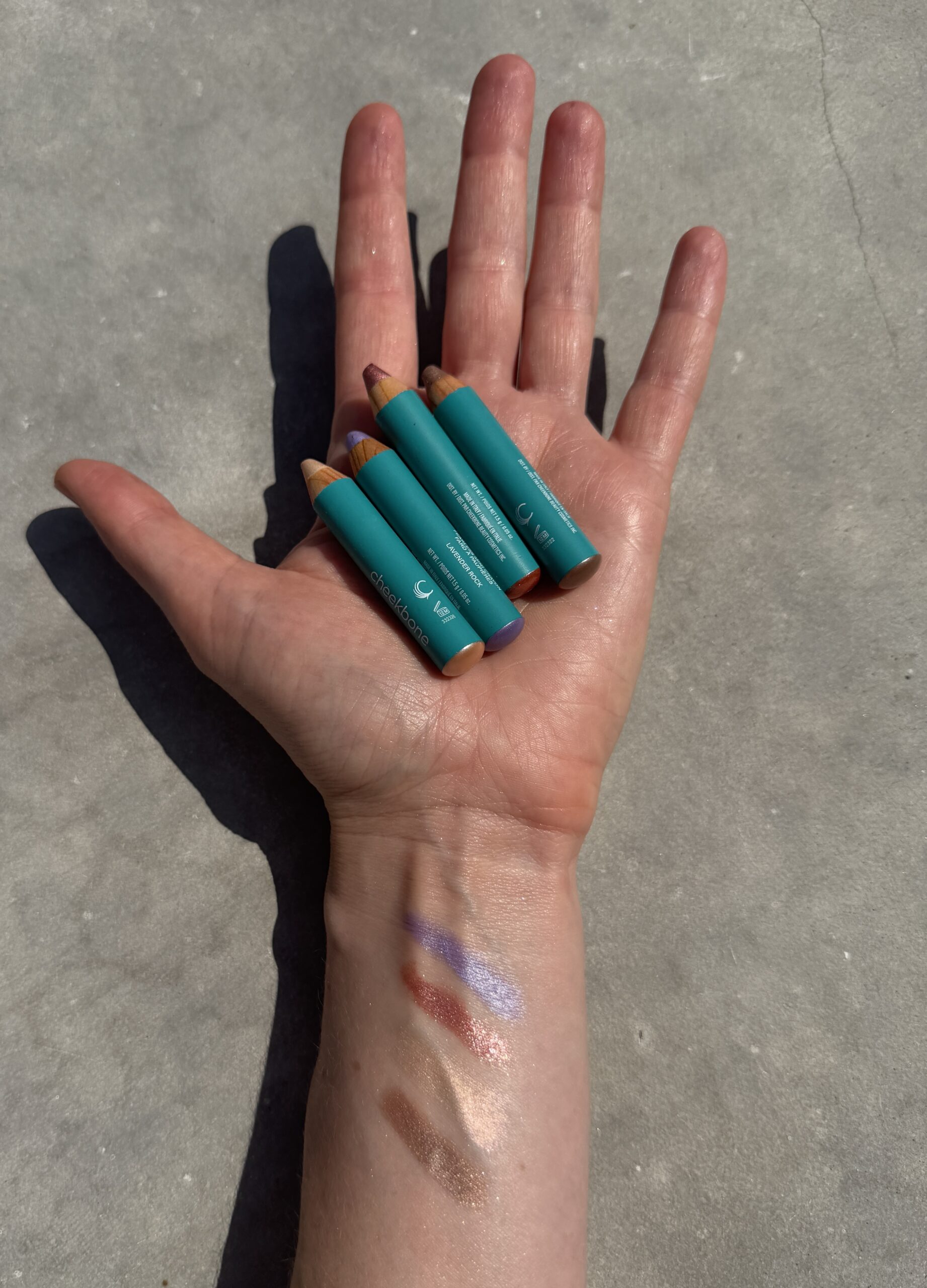 A hand holding four green makeup crayon sticks with swatches of their colors—brown, blue, bronze, and mauve—drawn on the forearm.
