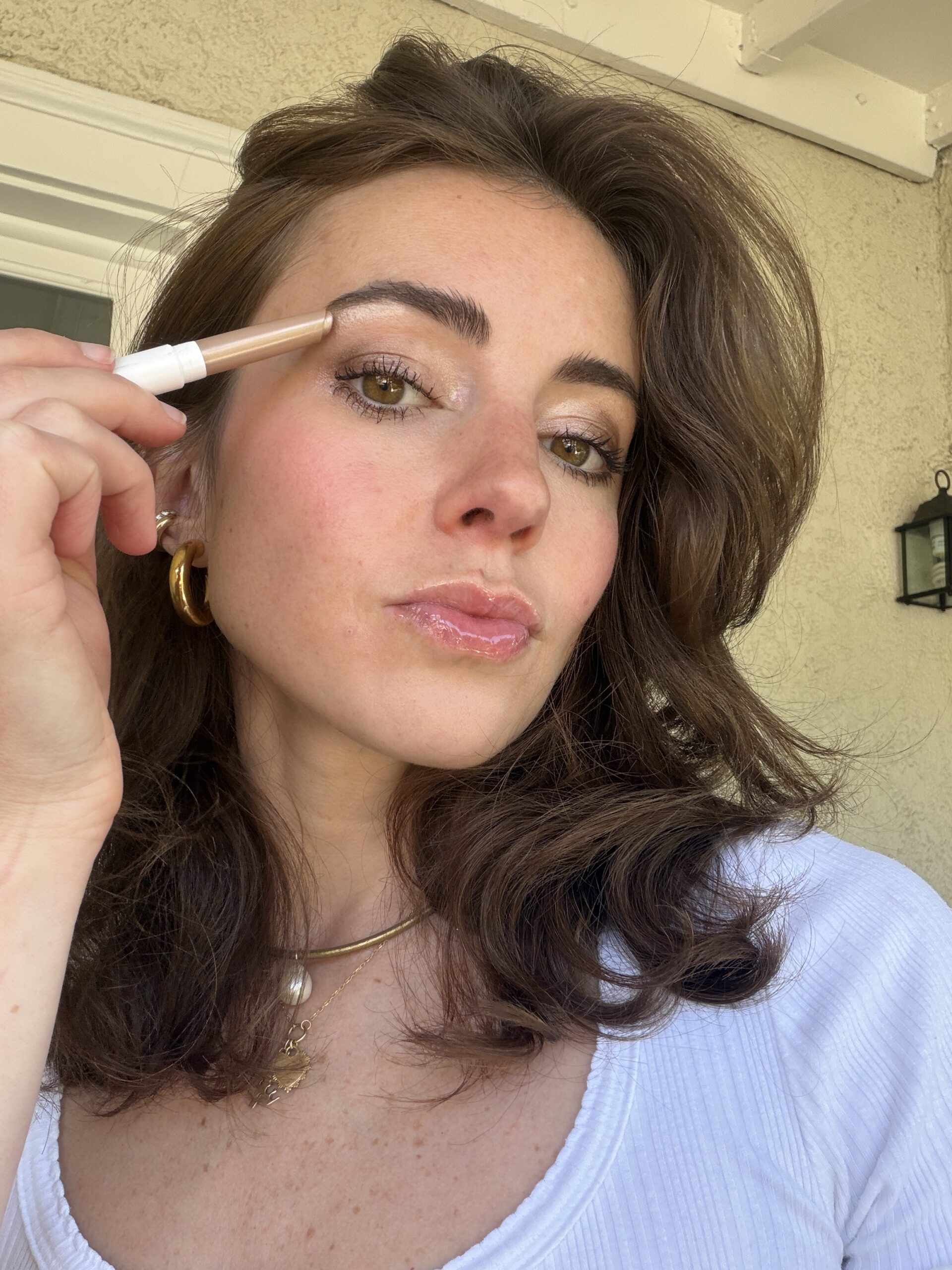 A woman with brown hair applies a makeup stick to her eyebrow while wearing a white top, gold jewelry, and glossy lip makeup.