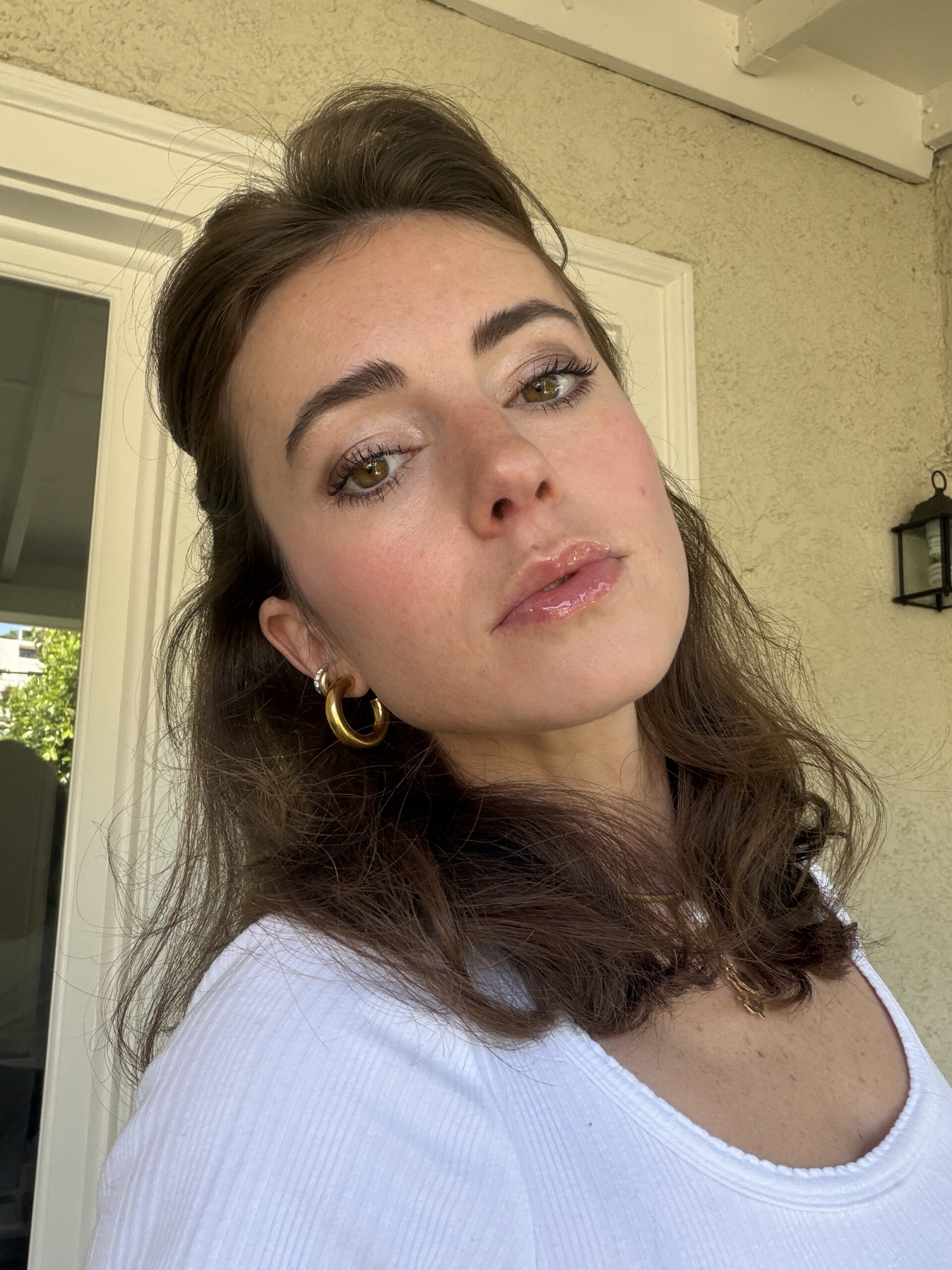 A woman with wavy brown hair and gold hoop earrings looks into the camera, wearing a white top, standing outside near a beige wall and a white door.