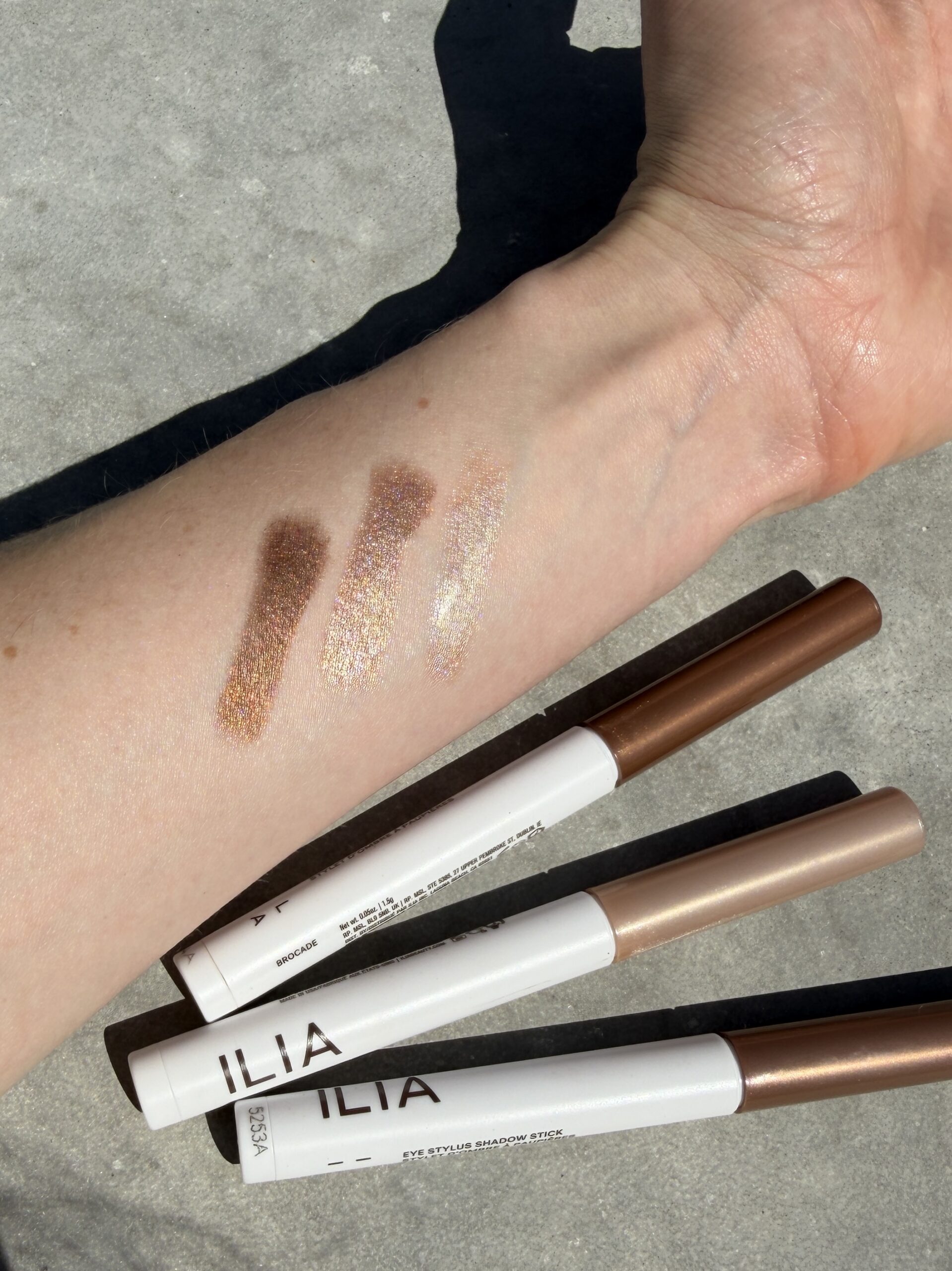 Three ILIA eyeshadow sticks are shown on a surface with matching swatches of shimmery brown and gold shades applied to a person's forearm.