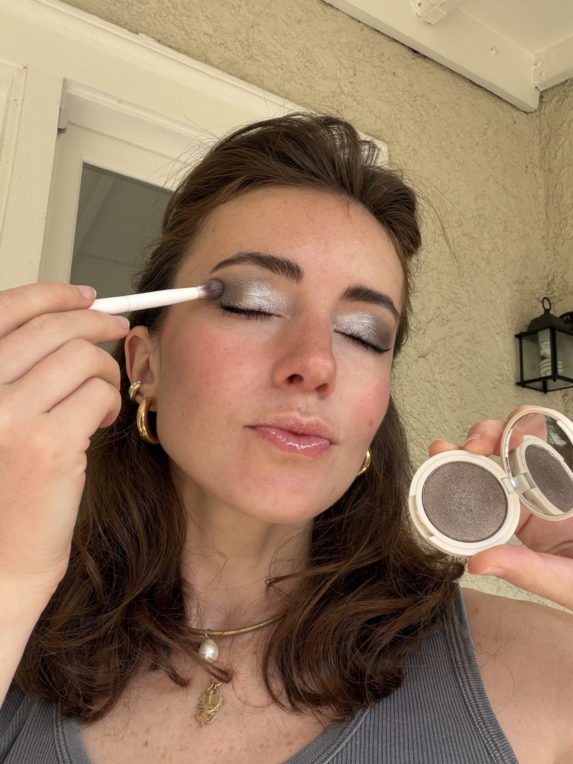 A woman applies silver eyeshadow to her eyelid with a brush while holding an open compact in her other hand.