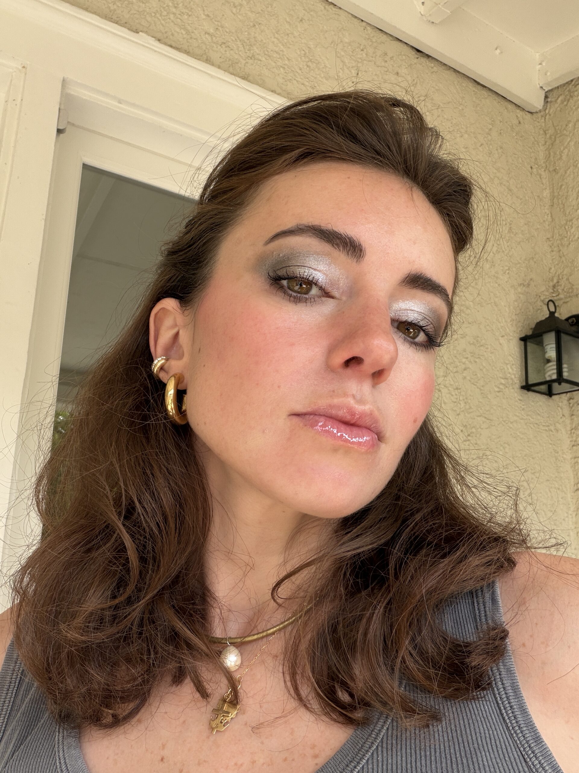 Woman with wavy brown hair wears hoop earrings, layered necklaces, and shimmery silver eye makeup, posing in front of a beige wall and door.