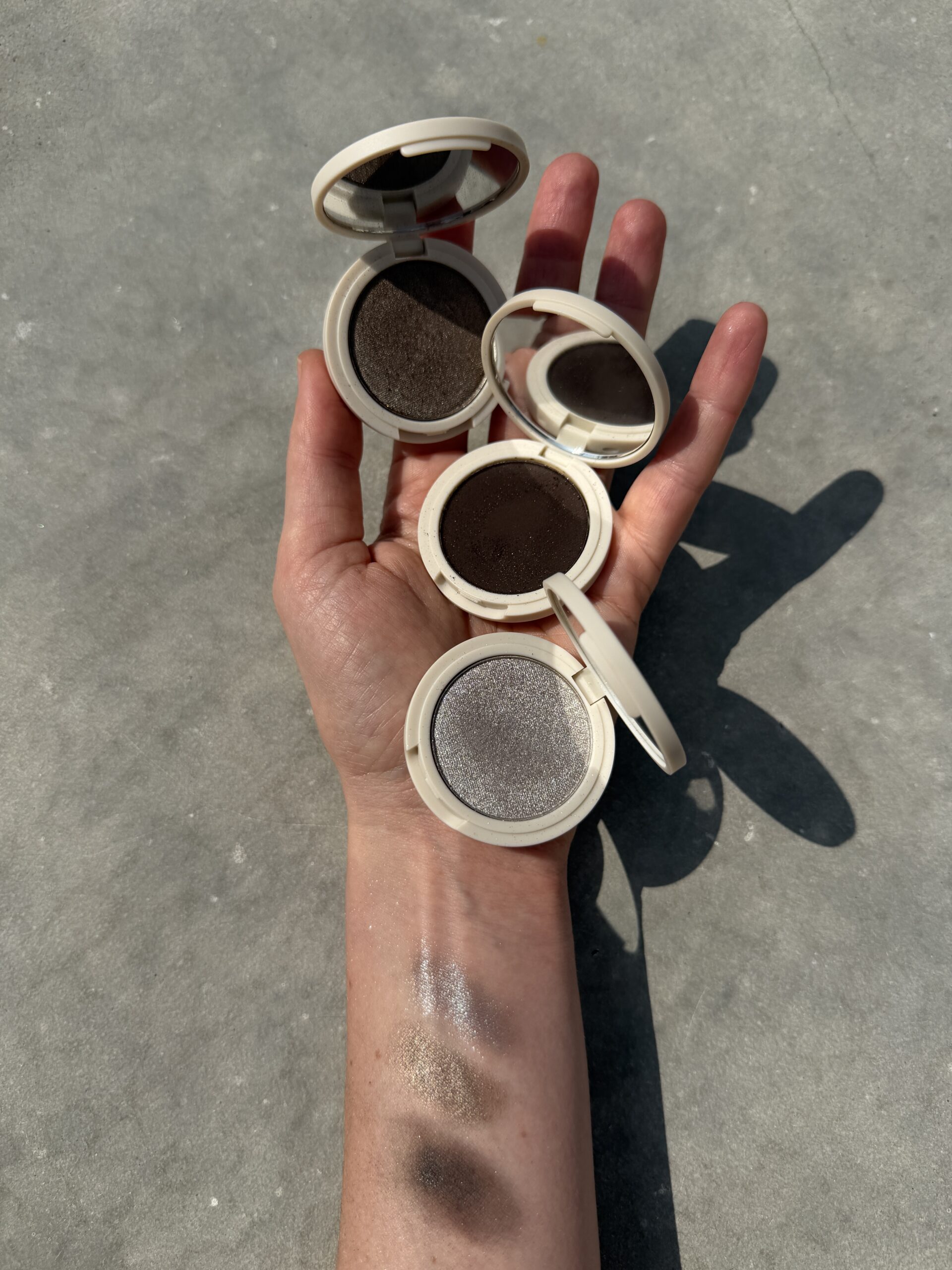 An open hand holds three round eyeshadow compacts in brown and gray shades, with swatches of the colors visible on the forearm.