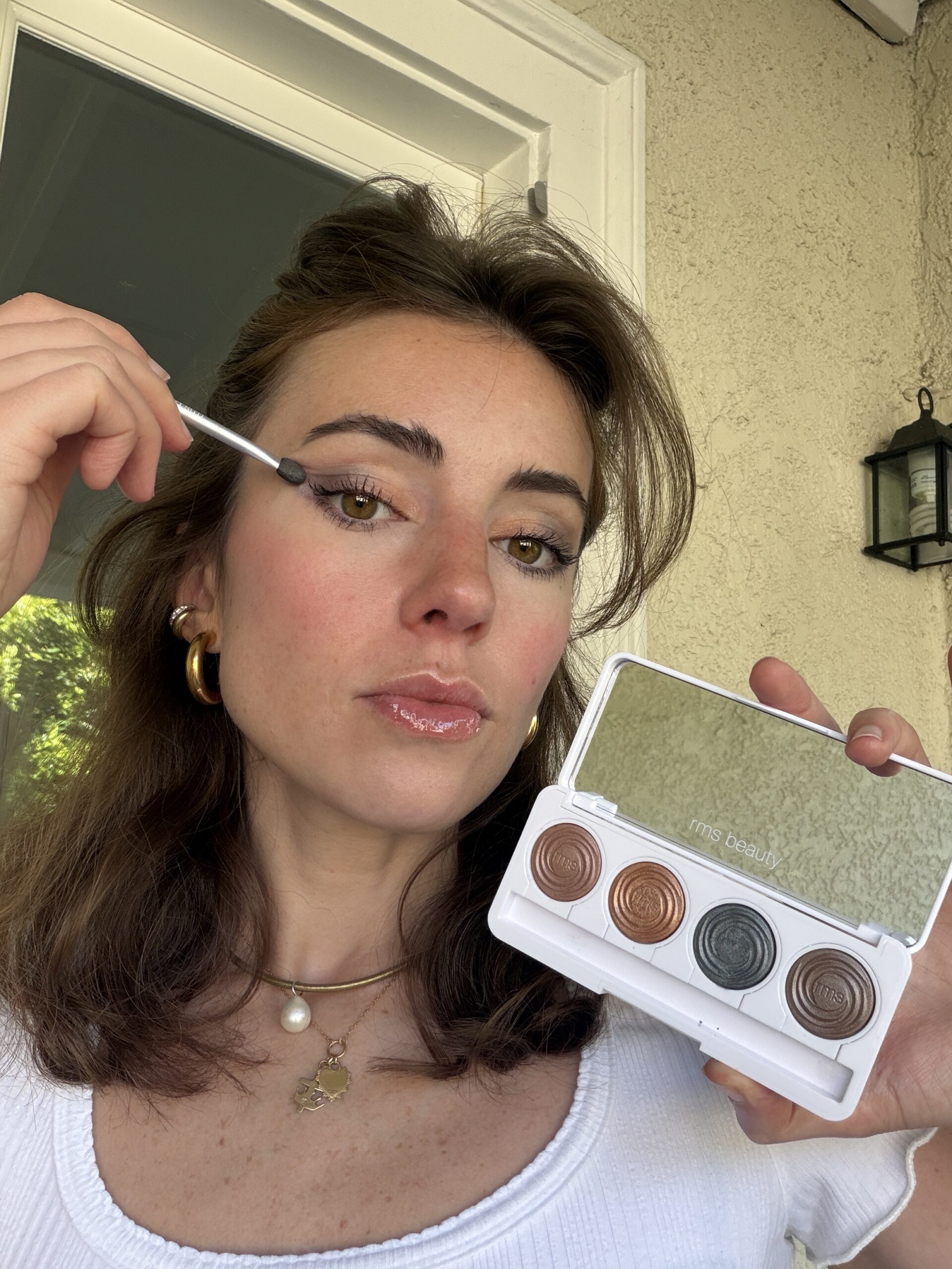 A woman applies eyeshadow with a brush, holding an open eyeshadow palette with four shades. She wears a white top, gold jewelry, and stands in front of a beige wall.