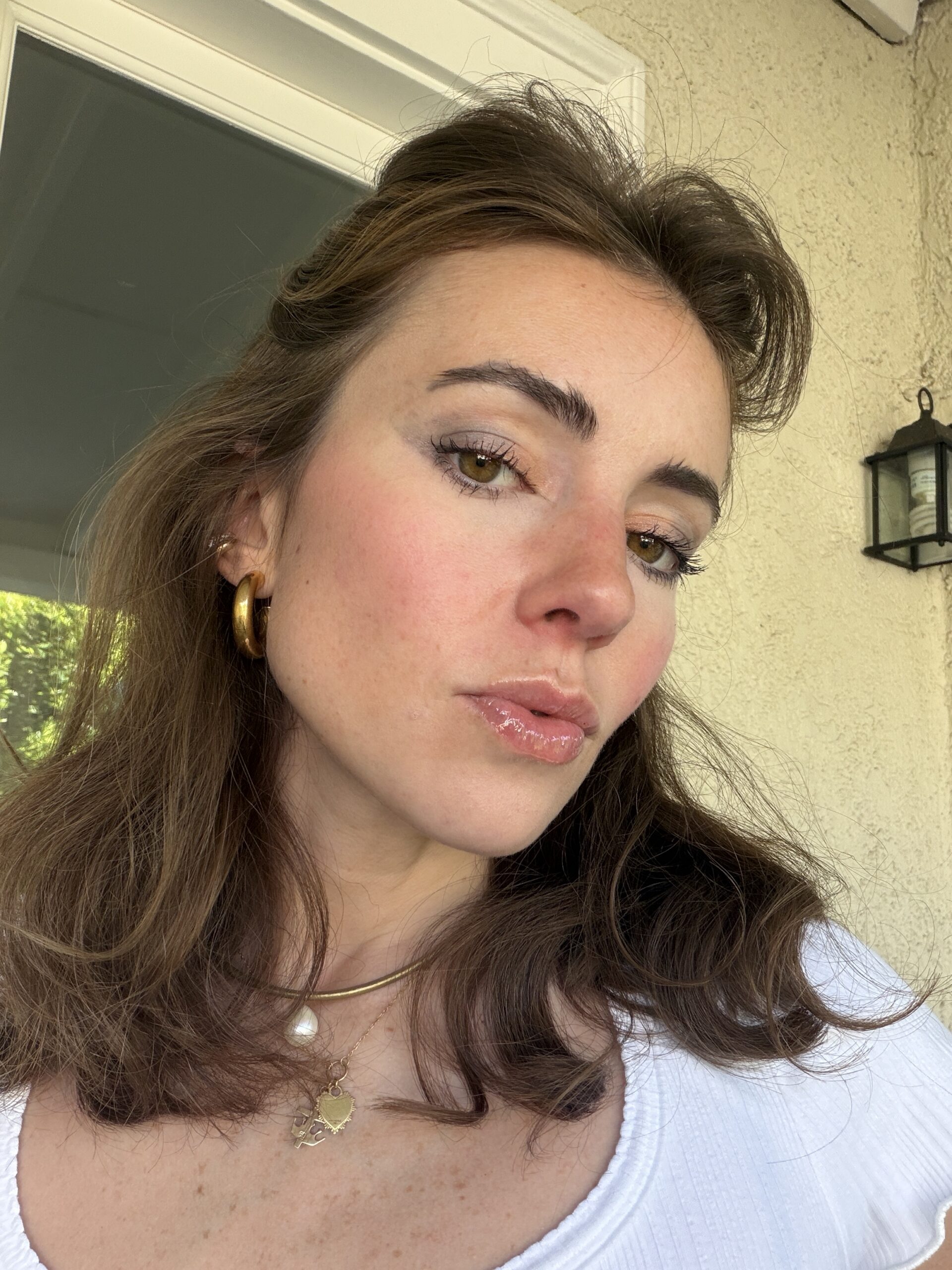 A woman with brown hair wearing a white top, gold hoop earring, layered necklaces, and natural makeup stands in front of a beige wall and window.