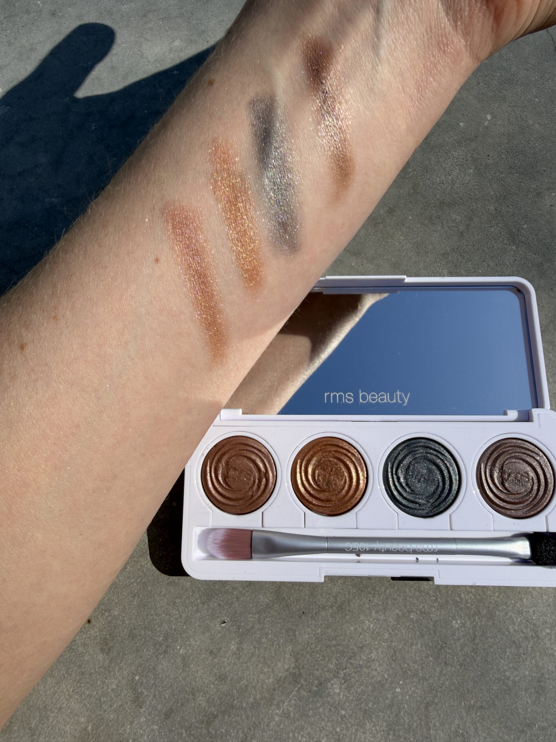 An arm displays swatches of four shimmery eyeshadow shades next to an open RMS Beauty palette with matching colors and a small brush.