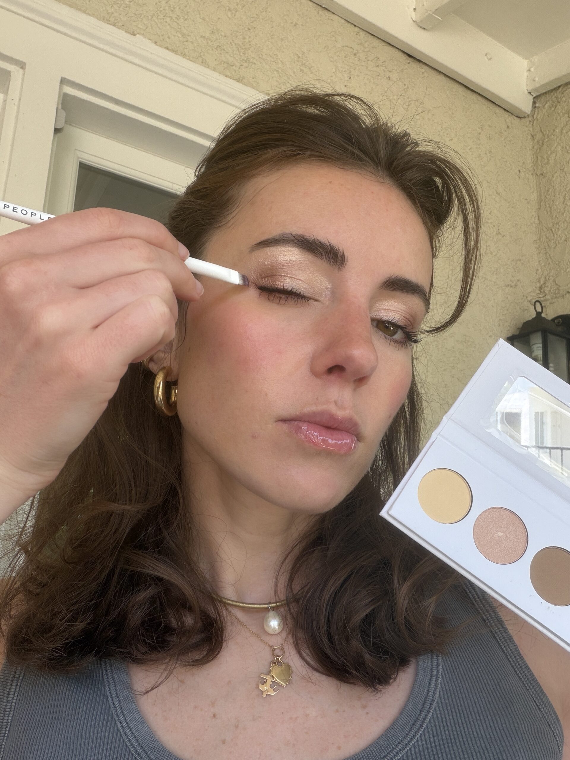 A woman applies shimmery eyeshadow to her eyelid using a brush, holding an eyeshadow palette with three shades in her other hand.