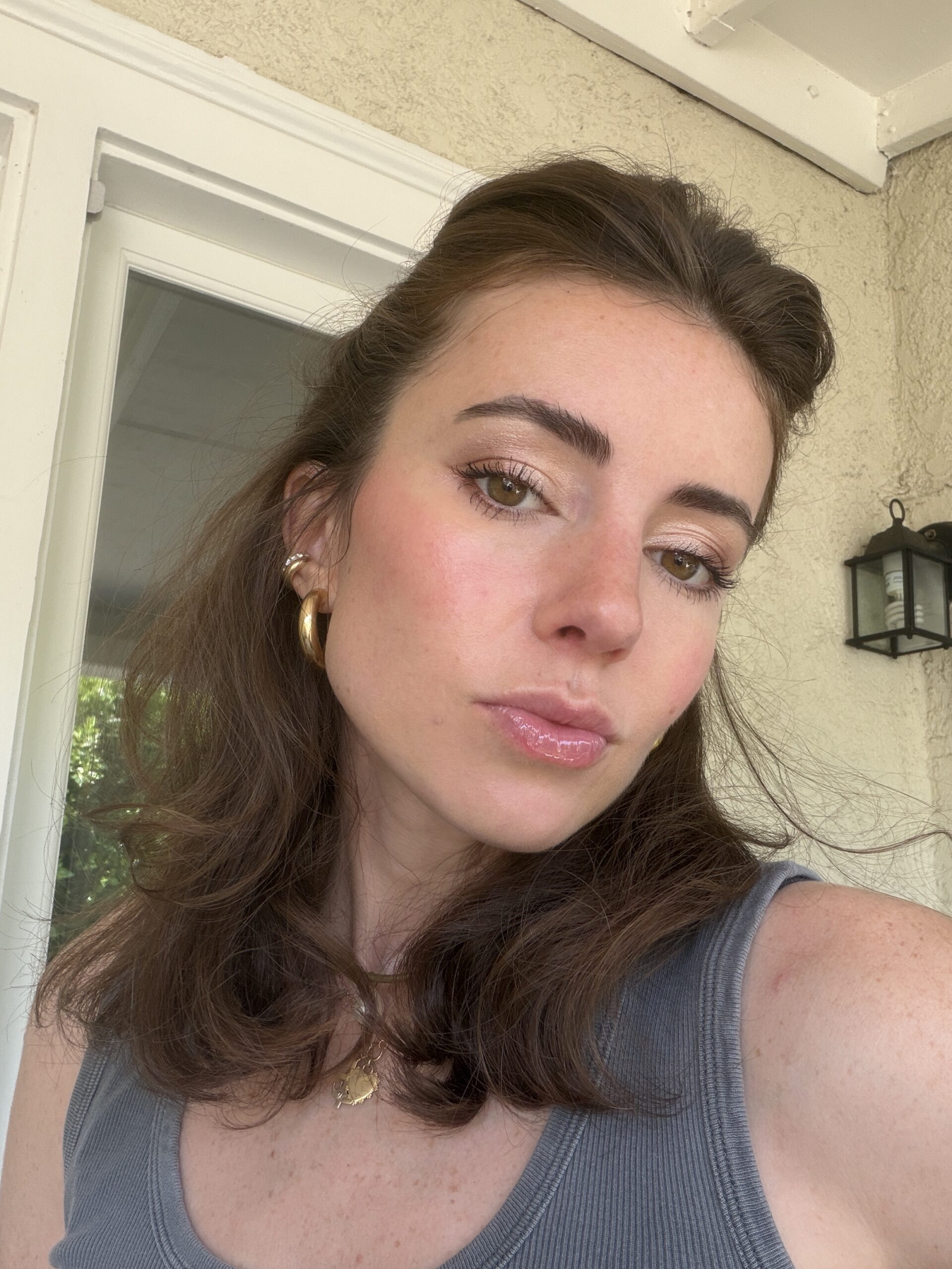 A woman with wavy brown hair, wearing a gray tank top and gold jewelry, takes a selfie outdoors in front of a light-colored wall and a glass door.