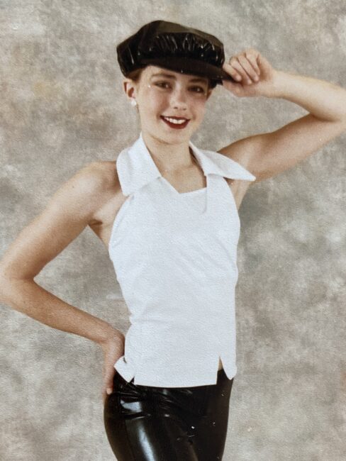 A person poses in a white sleeveless top, black shiny pants, and a black cap, smiling and tilting the cap with one hand against a neutral background.