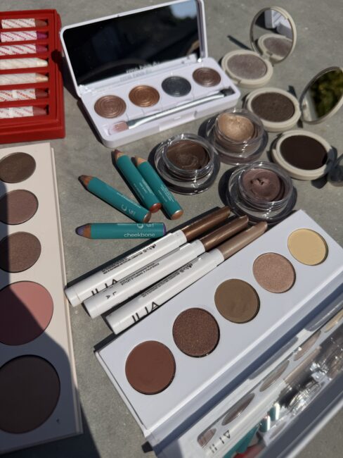 Various makeup products, including eyeshadow palettes, cream eyeshadows, brow pencils, and crayon sticks, are arranged on a gray surface in natural daylight.