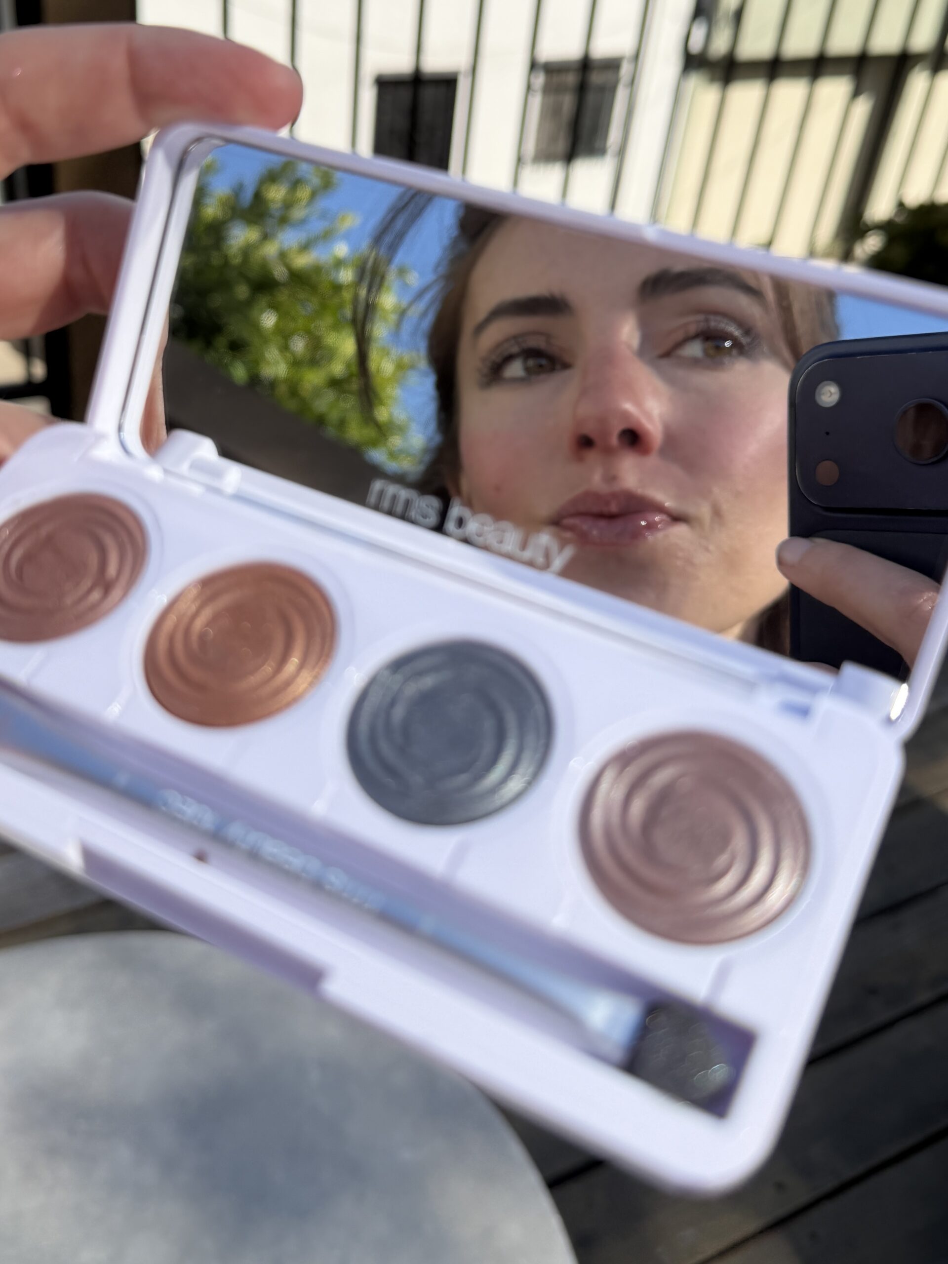 A person takes a selfie using a smartphone, capturing their reflection in a makeup palette mirror with four eyeshadow shades and a brush.