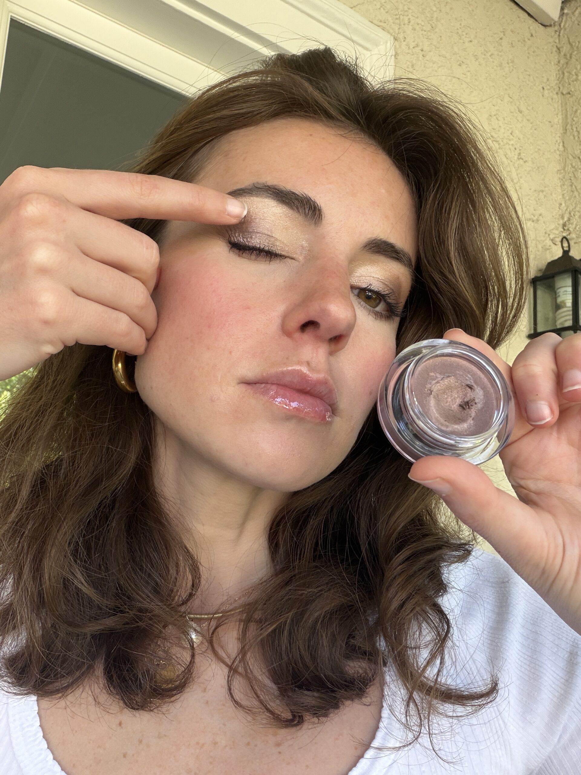 Woman applying shimmery eyeshadow to her eyelid with one hand while holding an open jar of the eyeshadow with the other hand.