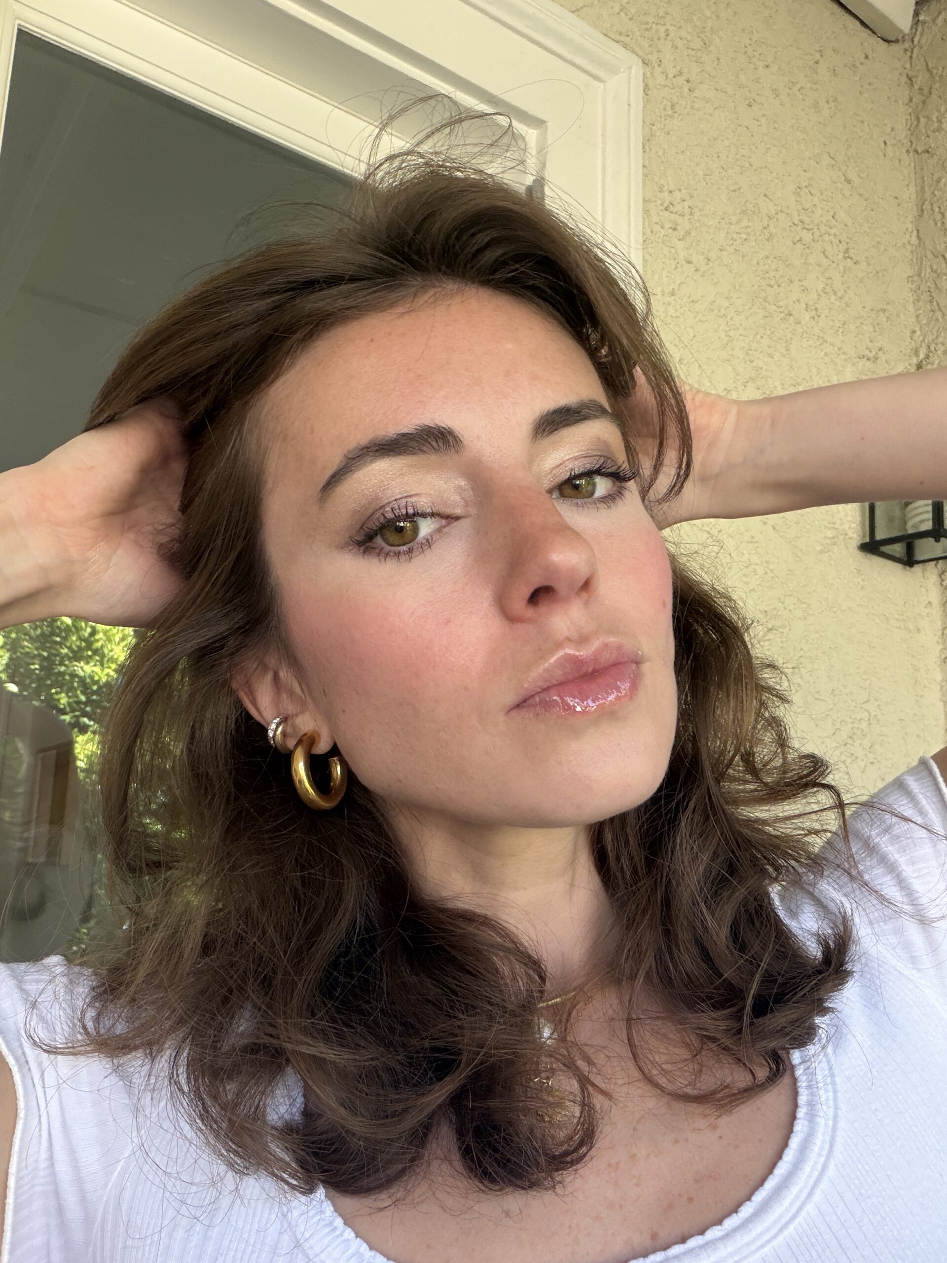 Woman with wavy brown hair wearing a white top and gold hoop earrings poses with hands in her hair in front of a beige wall and glass door.