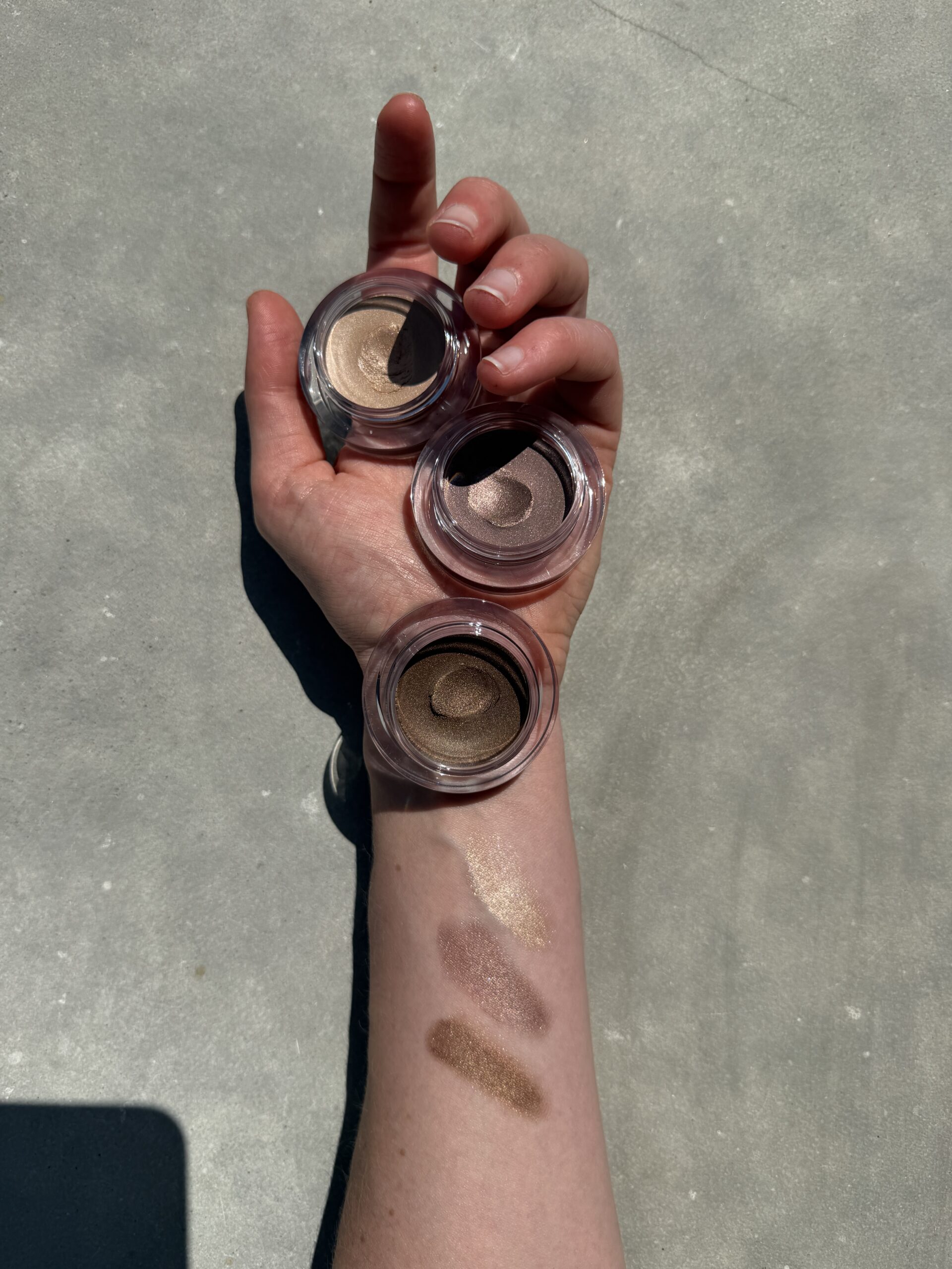 A hand holds three open eyeshadow pots in neutral shades, with corresponding swatches displayed on the forearm against a plain background.