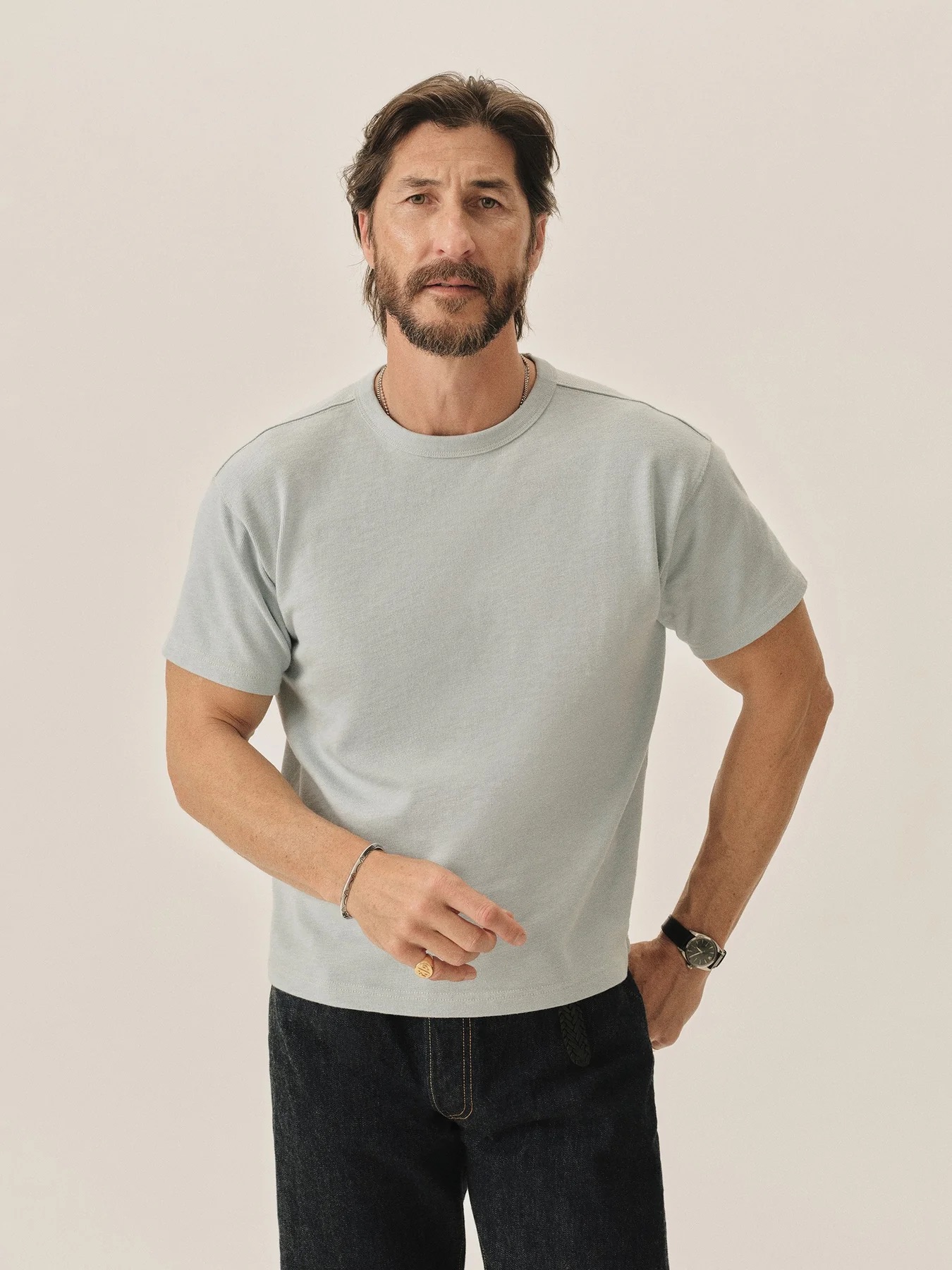 A man with dark hair and a beard wearing a light gray t-shirt and dark jeans stands against a plain, light background.