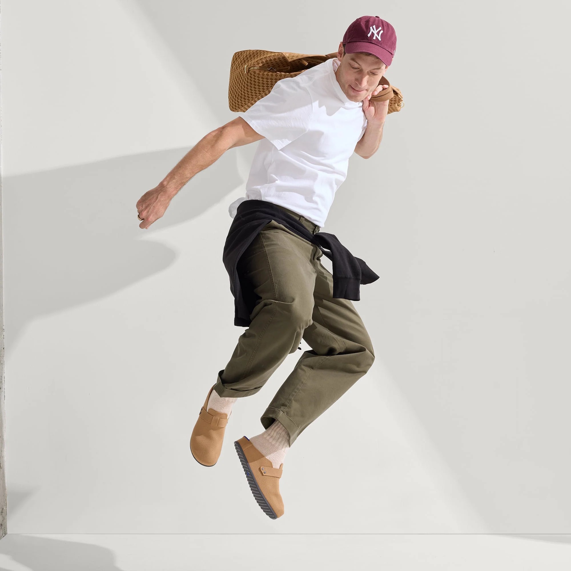 A man wearing a maroon baseball cap, white t-shirt, olive pants, tan shoes, and carrying a woven bag jumps in front of a plain light background.