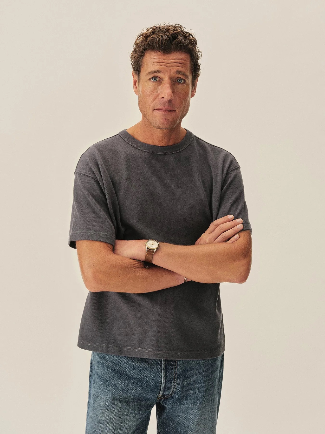 Man standing with arms crossed, wearing a dark gray t-shirt, blue jeans, and a wristwatch, looking directly at the camera against a plain light background.