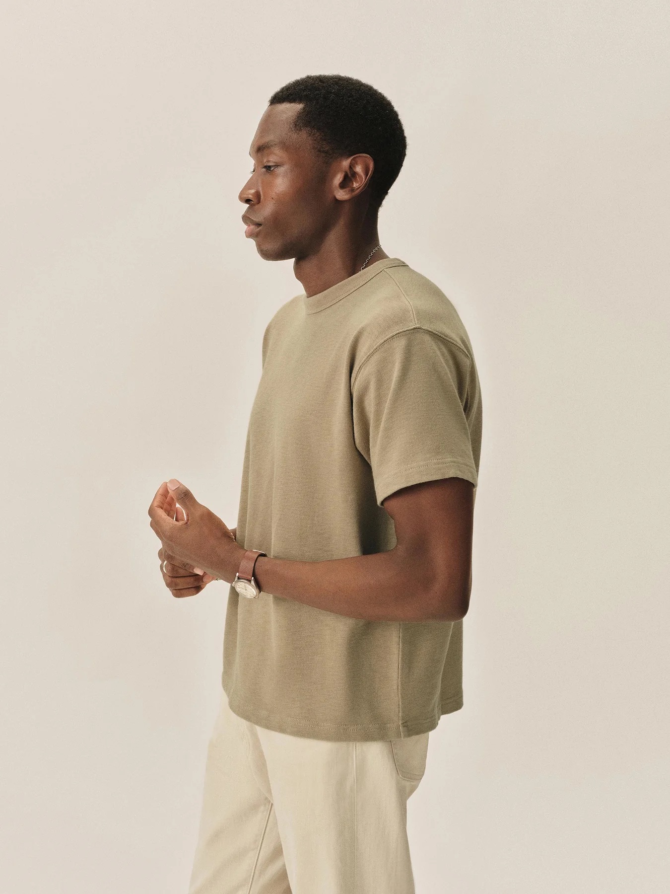 A man stands in profile against a plain background, wearing a beige short-sleeve t-shirt and light-colored pants.