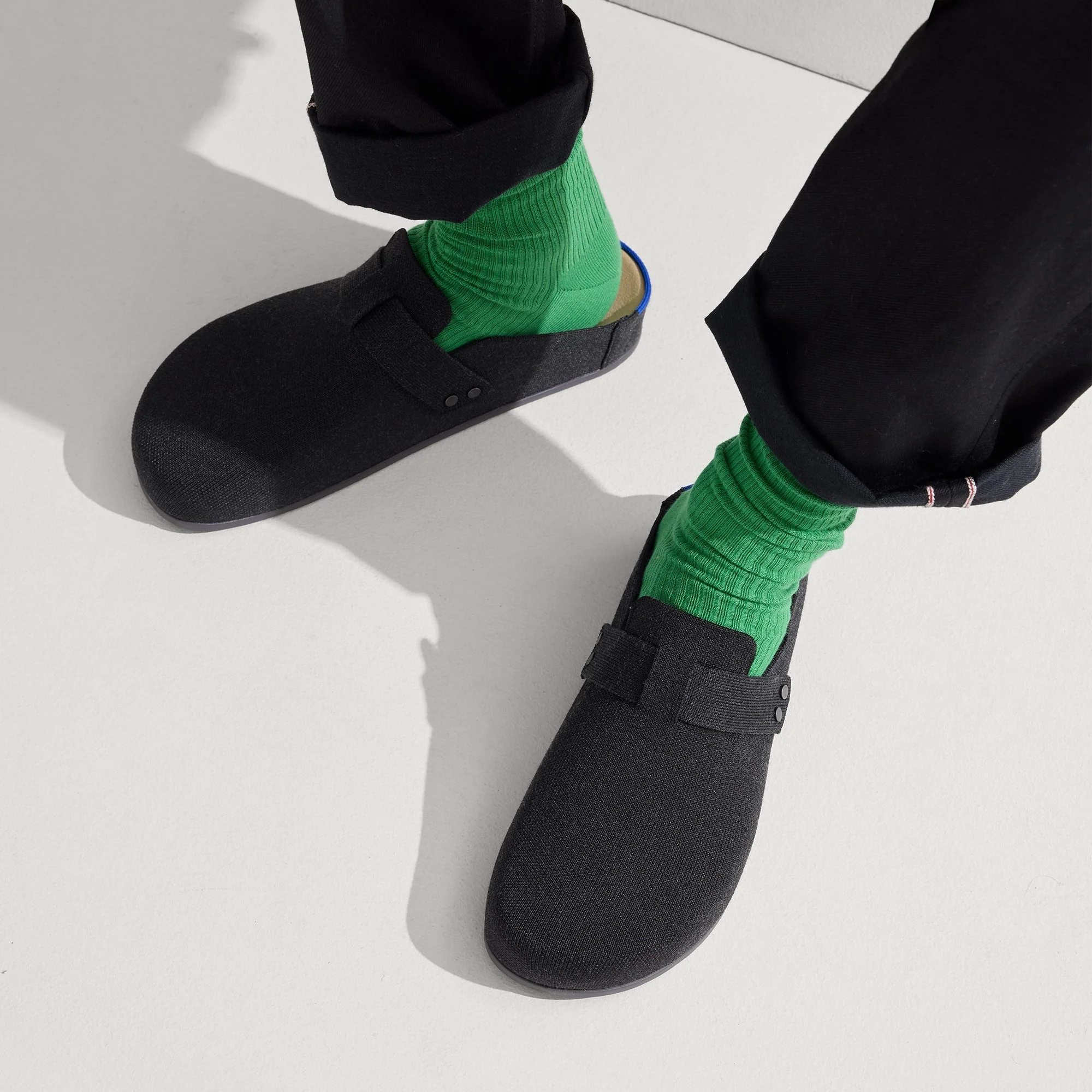 Person wearing bright green socks and black slip-on shoes, standing on a light-colored floor, with black pants slightly rolled up at the ankle.