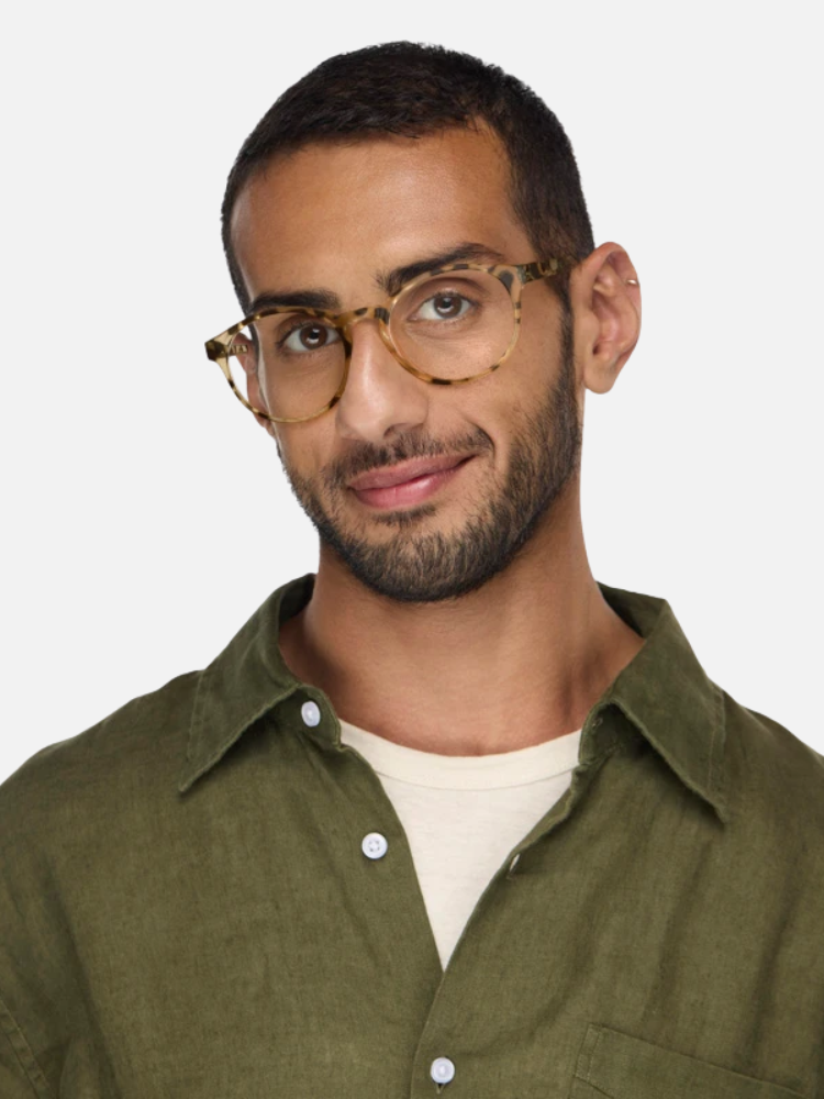 Man with short dark hair, beard, and tortoise-shell glasses wearing an olive green button-up shirt over a cream t-shirt, posed against a plain light background.