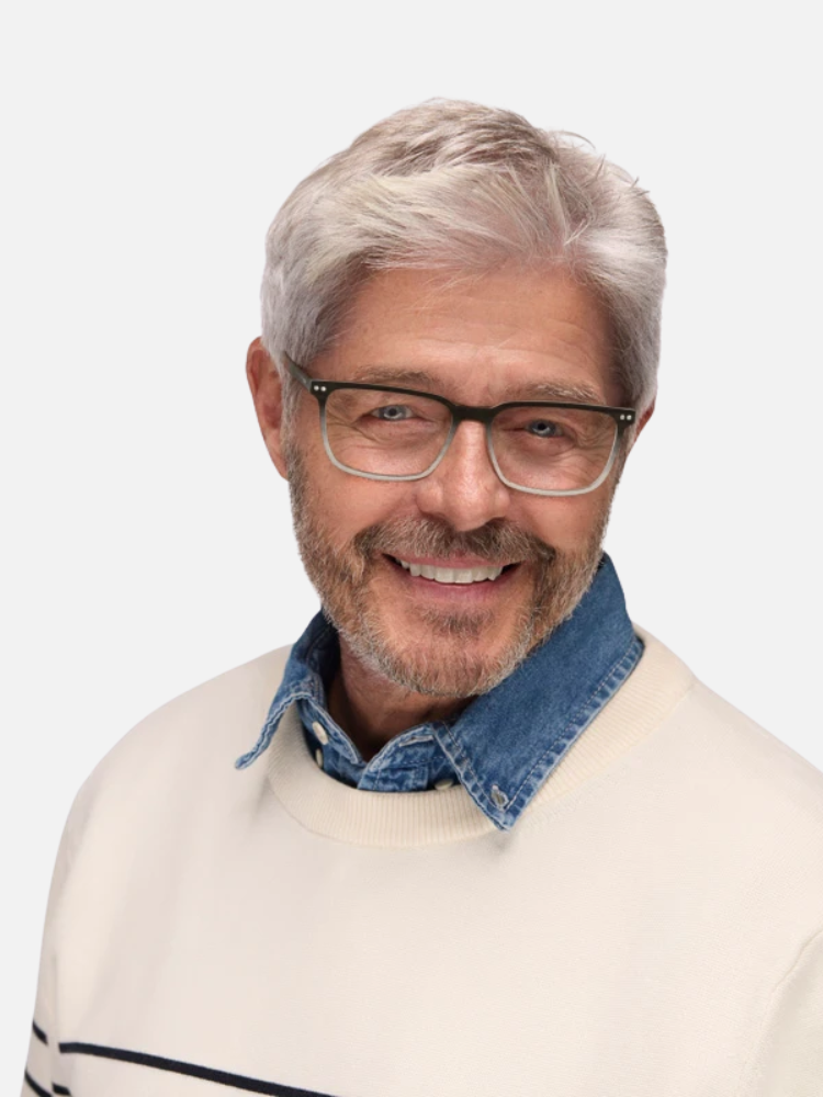 Smiling older man with gray hair, beard, and glasses, wearing a light sweater over a denim collared shirt, posed against a plain light background.